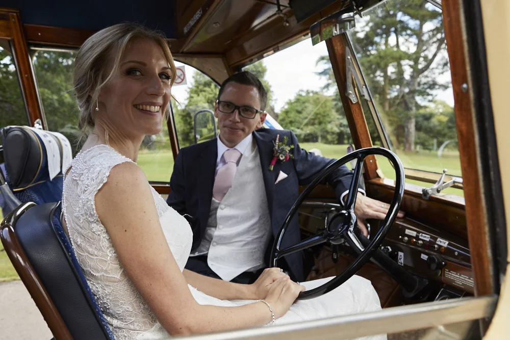 A bride and groom sitting in a vintage car during their wedding day. The bride is smiling and wearing a lace wedding dress, while the groom is dressed in a dark suit with a boutonniere, sitting next to her at the steering wheel. The background shows trees and greenery.