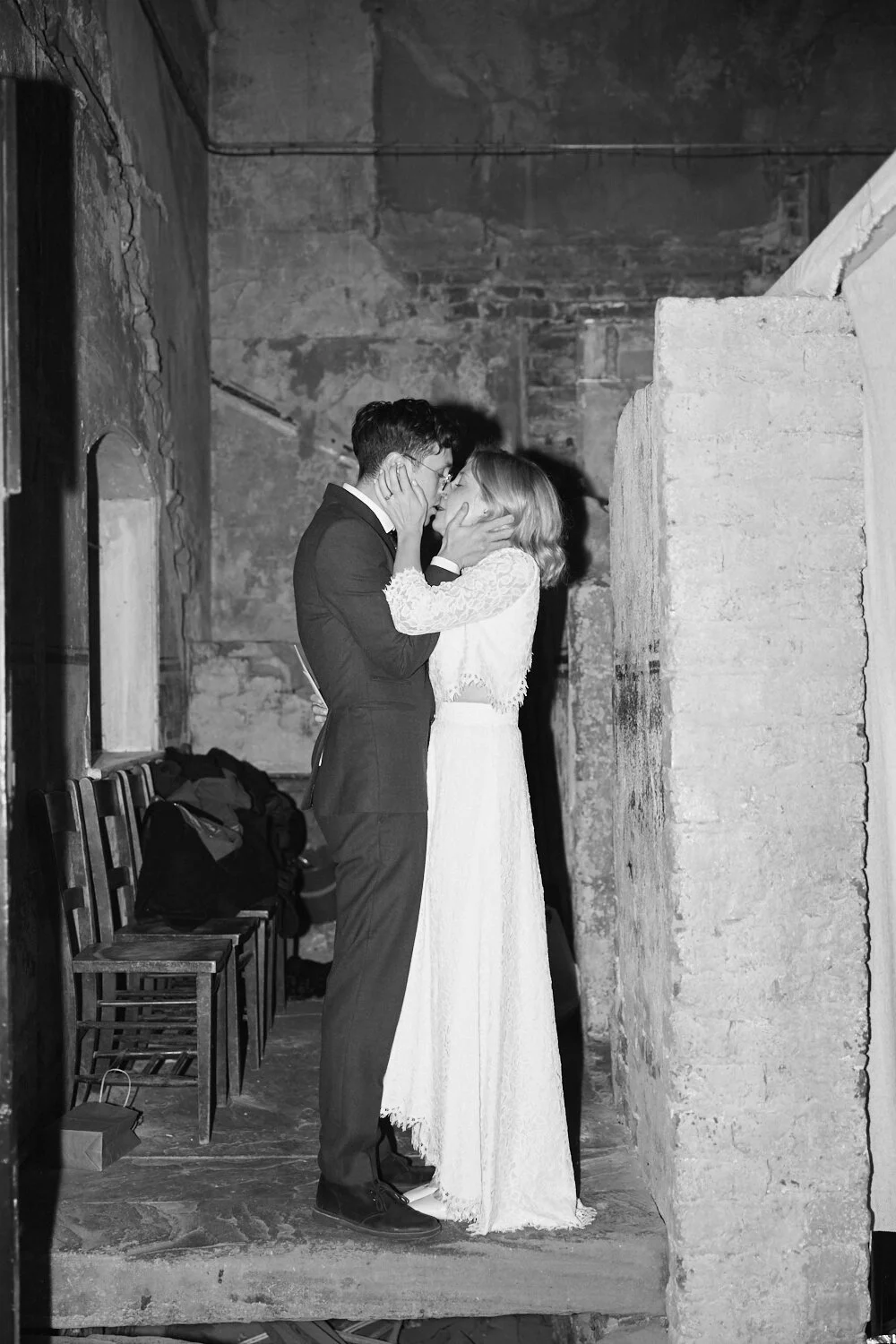 A black and white photo of a couple embracing and kissing in an intimate moment inside a rustic, industrial space with exposed brick walls and concrete. The woman wears a lace dress, and the man is in a suit.