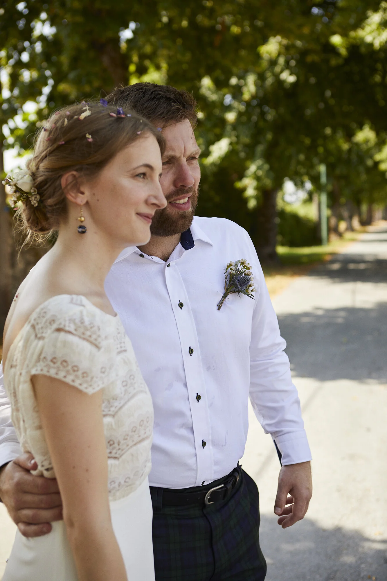 A woman and a man standing outdoors on a sunny day, with trees in the background. The woman has brown hair styled in a bun with small flowers and is wearing a white lace dress. The man has a beard, is wearing a white shirt with a flower boutonniere, 