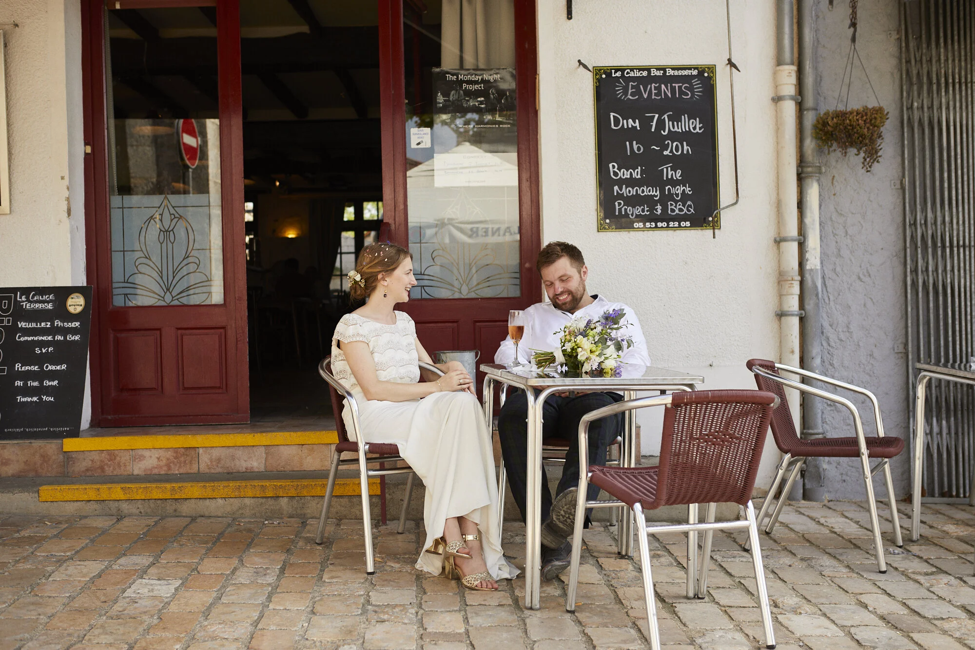 A man and woman sitting at an outdoor table of a French brasserie, smiling and enjoying each other's company, with a bouquet of flowers on the table and a glass of rosé wine.