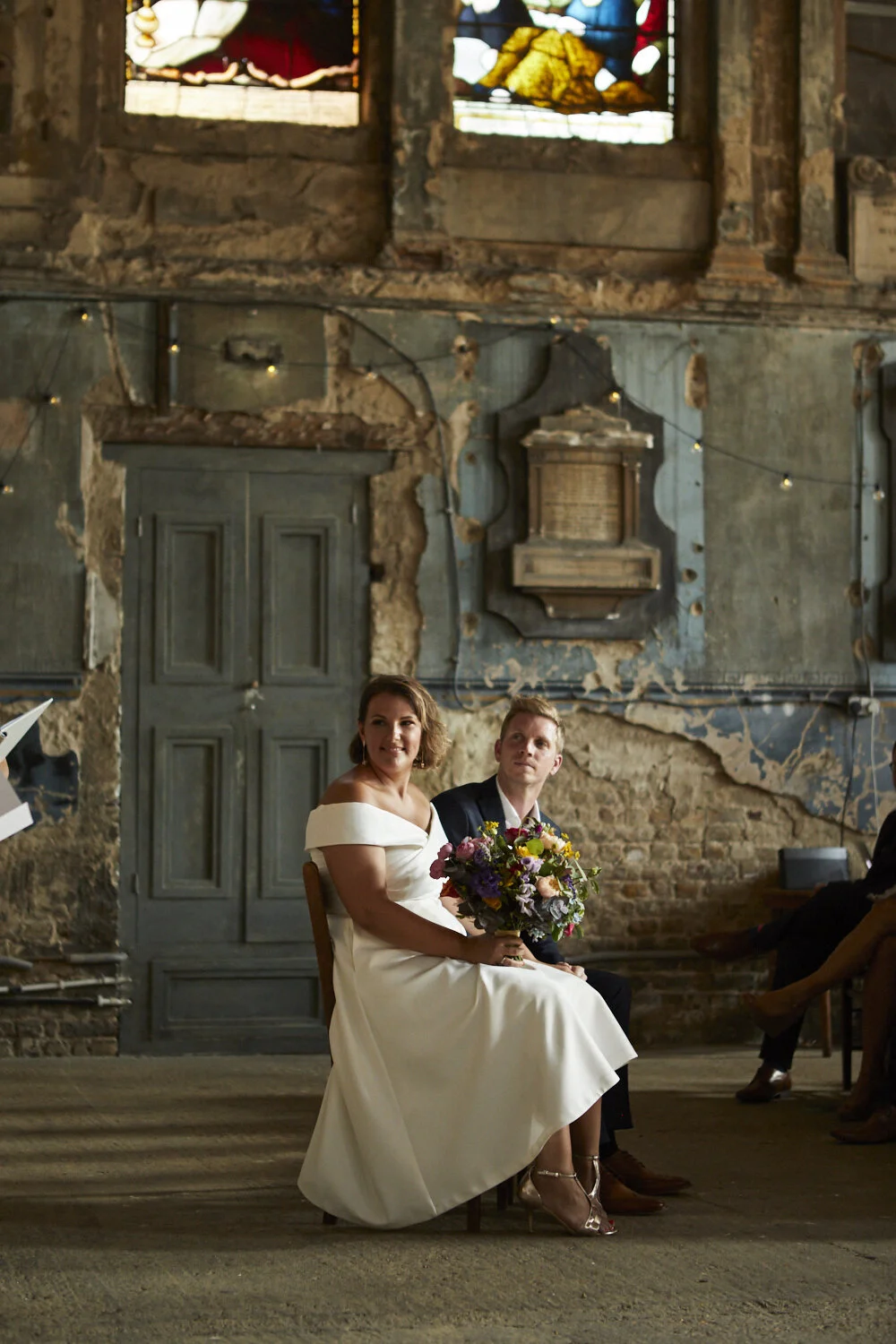 A bride in a white wedding dress and a groom in a dark suit sit together during a wedding ceremony inside an industrial-style venue with exposed brick walls and stained glass windows. The bride holds a colorful bouquet of flowers, and both are looking slightly to the side.