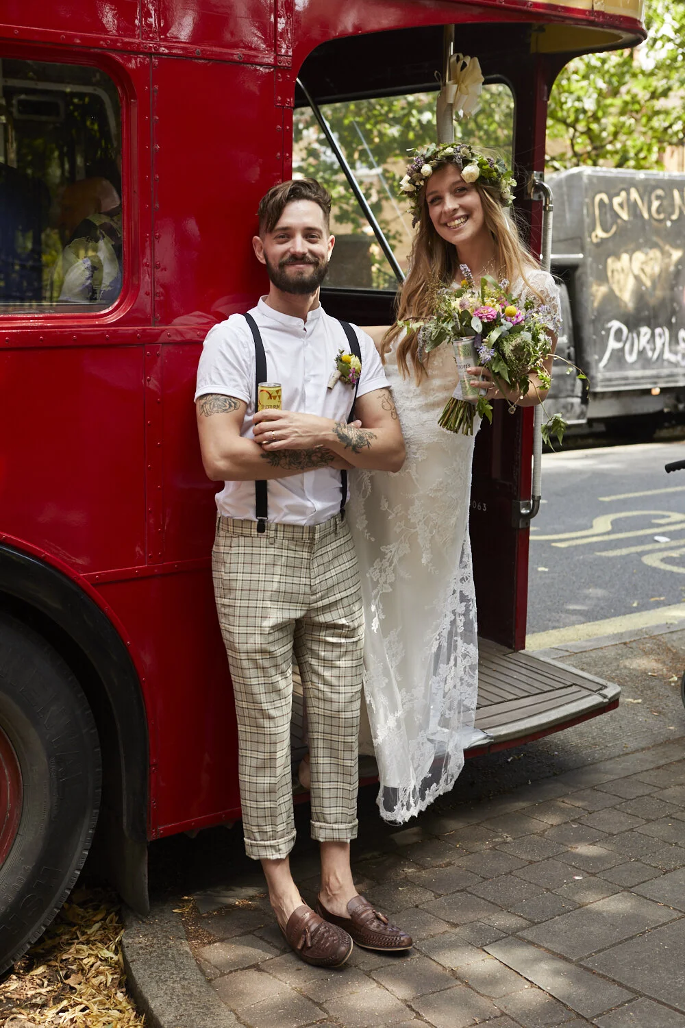 A man and woman in wedding attire standing next to a vintage red truck. The woman is holding a bouquet and wearing a floral crown, while the man is drinking from a can and has tattoos and suspenders.