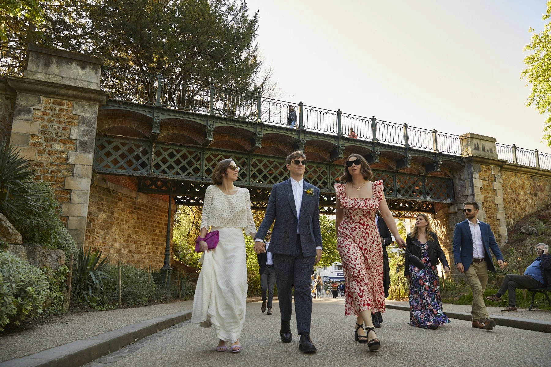 People dressed in formal attire walking under a bridge in a park surrounded by trees and greenery.