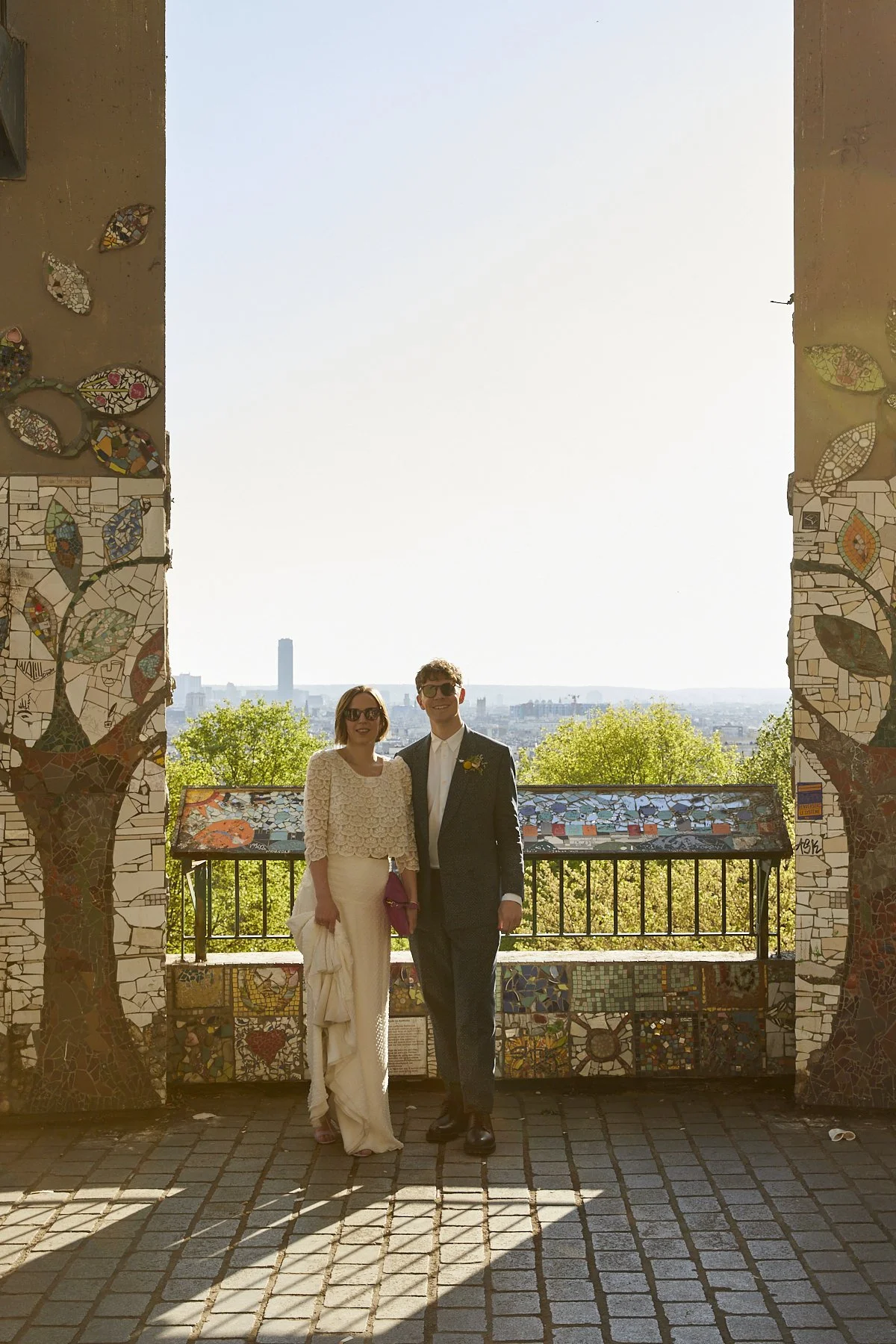 A couple dressed in formal attire standing on a balcony with a mosaic mural, overlooking a city skyline with tall buildings and trees, during daytime.