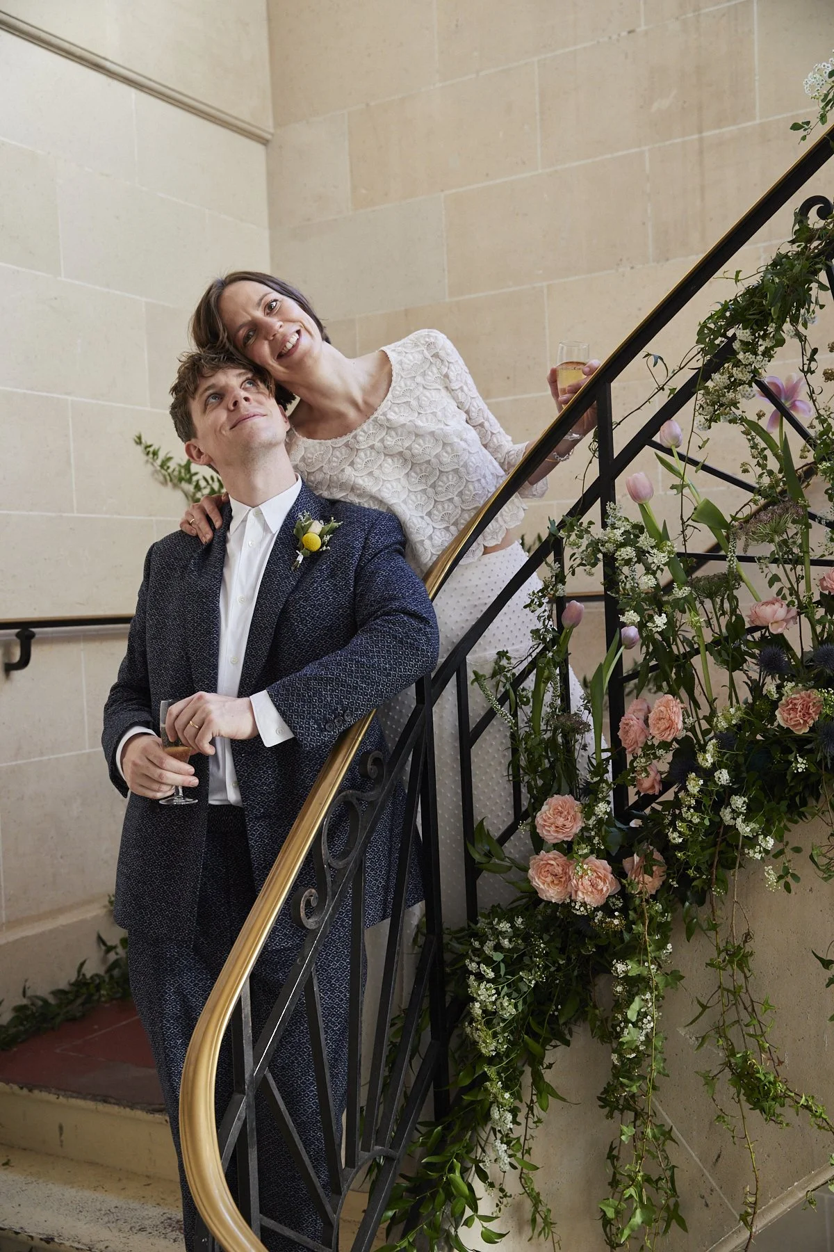 A couple standing on a staircase with floral decorations, celebrating with drinks. The man is in a blue suit with a white shirt and a boutonniere, looking up at the woman, who is wearing a lace dress and holding a glass of champagne, smiling.