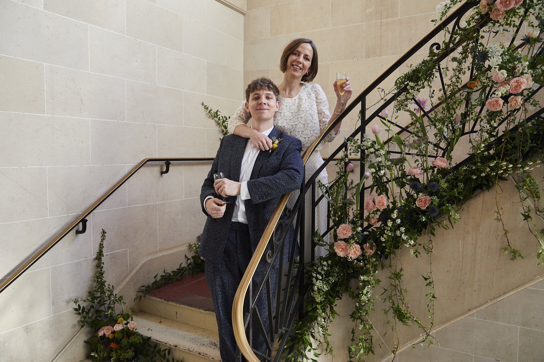 A man in a suit and a woman in a white dress celebrating with champagne on a decorated staircase.