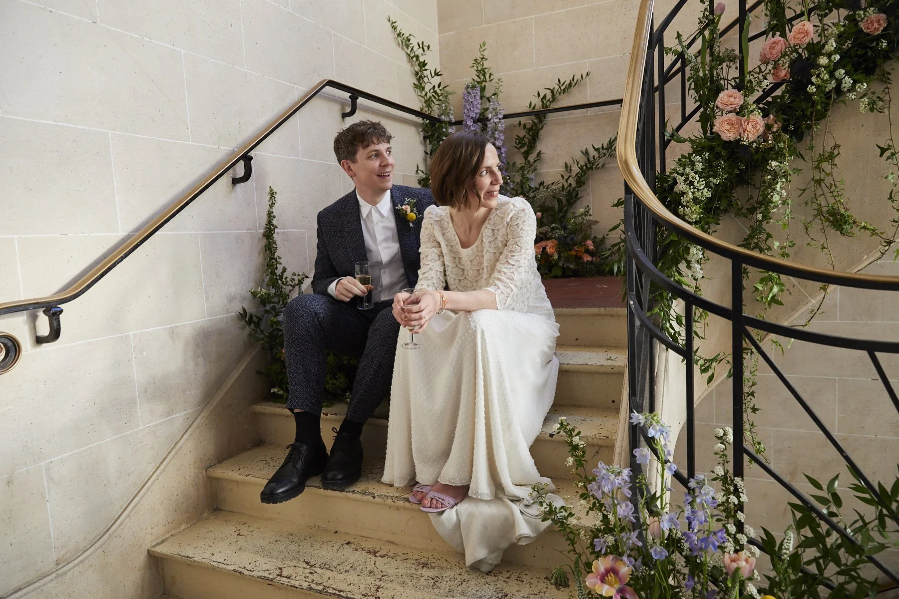 A bride and groom sit on a staircase decorated with flowers, holding glasses, during their wedding celebration.