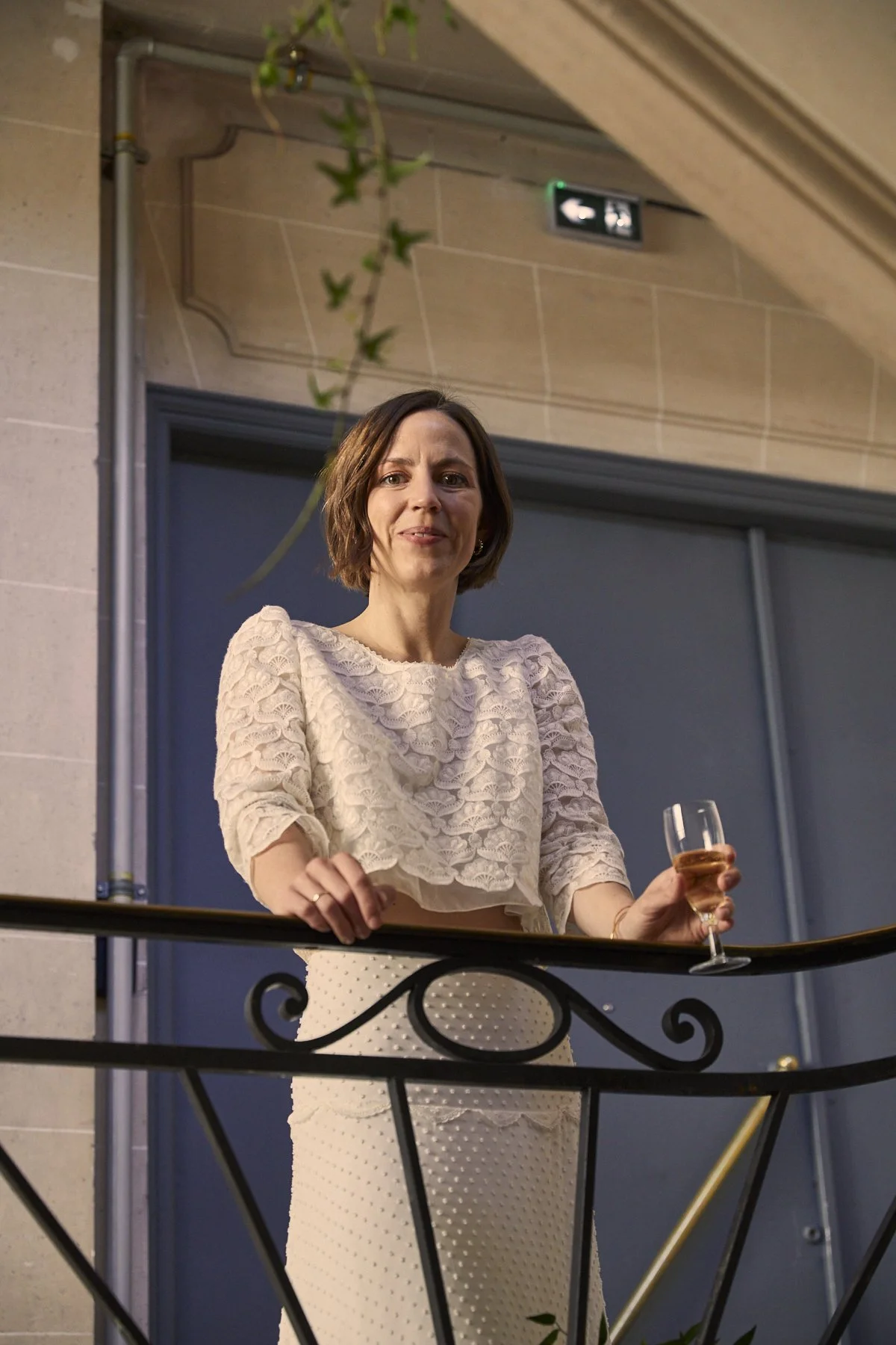 A woman with short brown hair standing on a balcony, holding a glass of champagne, smiling and looking at the camera. She is wearing a cream-colored lace top and a polka dot skirt, with a decorative wrought iron railing in front of her.