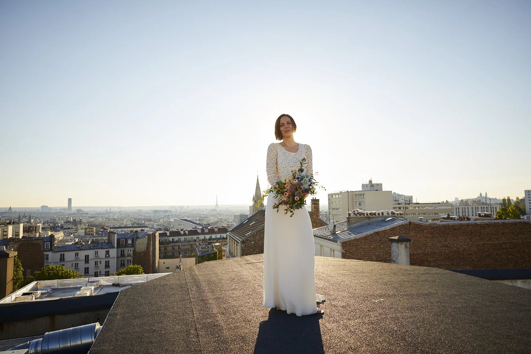 A woman in a white dress with lace sleeves holding a bouquet of flowers stands on a rooftop with a cityscape in the background during sunset.