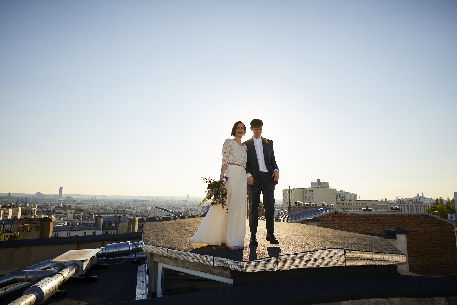 A newlywed couple standing on a rooftop at sunset with the city skyline in the background, holding hands and smiling.