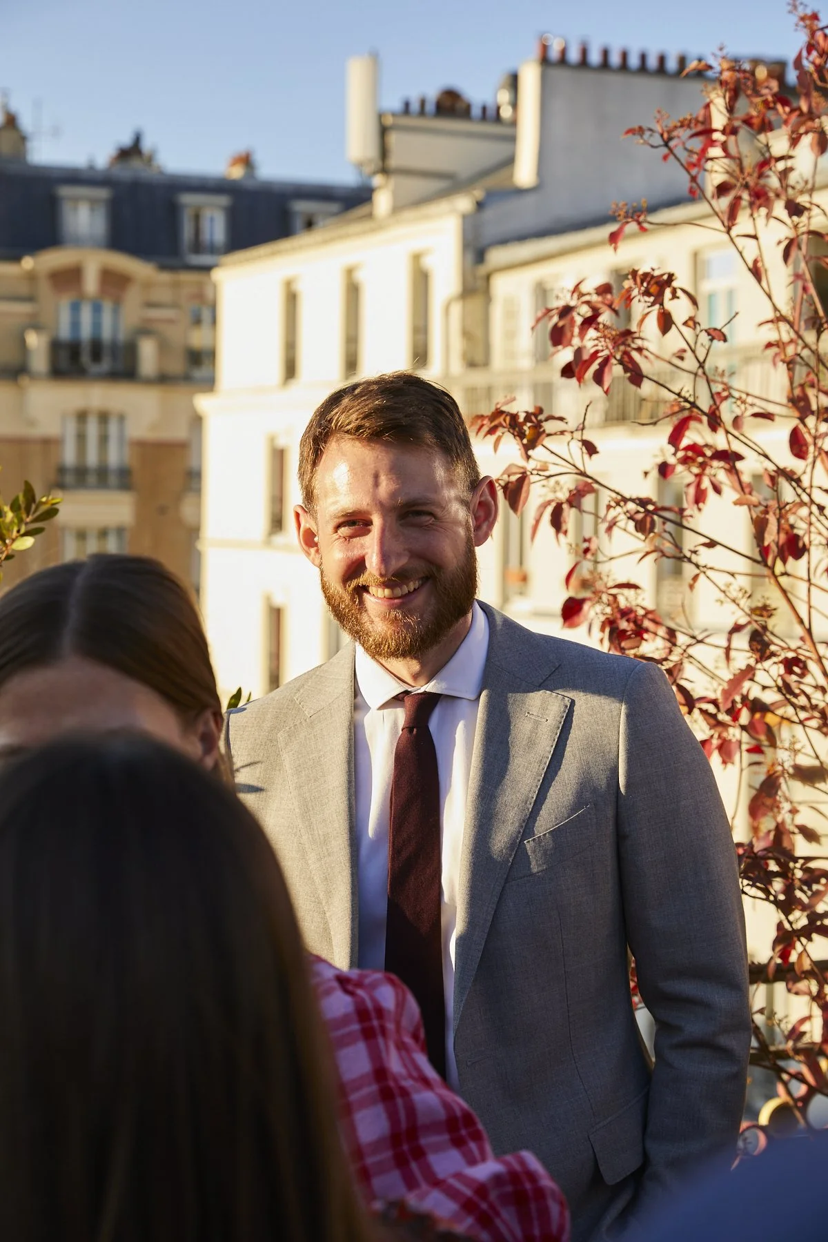 A man with a beard smiling in a gray suit and tie, standing outside during sunset, with red-leaved trees and Parisian-style buildings in the background.