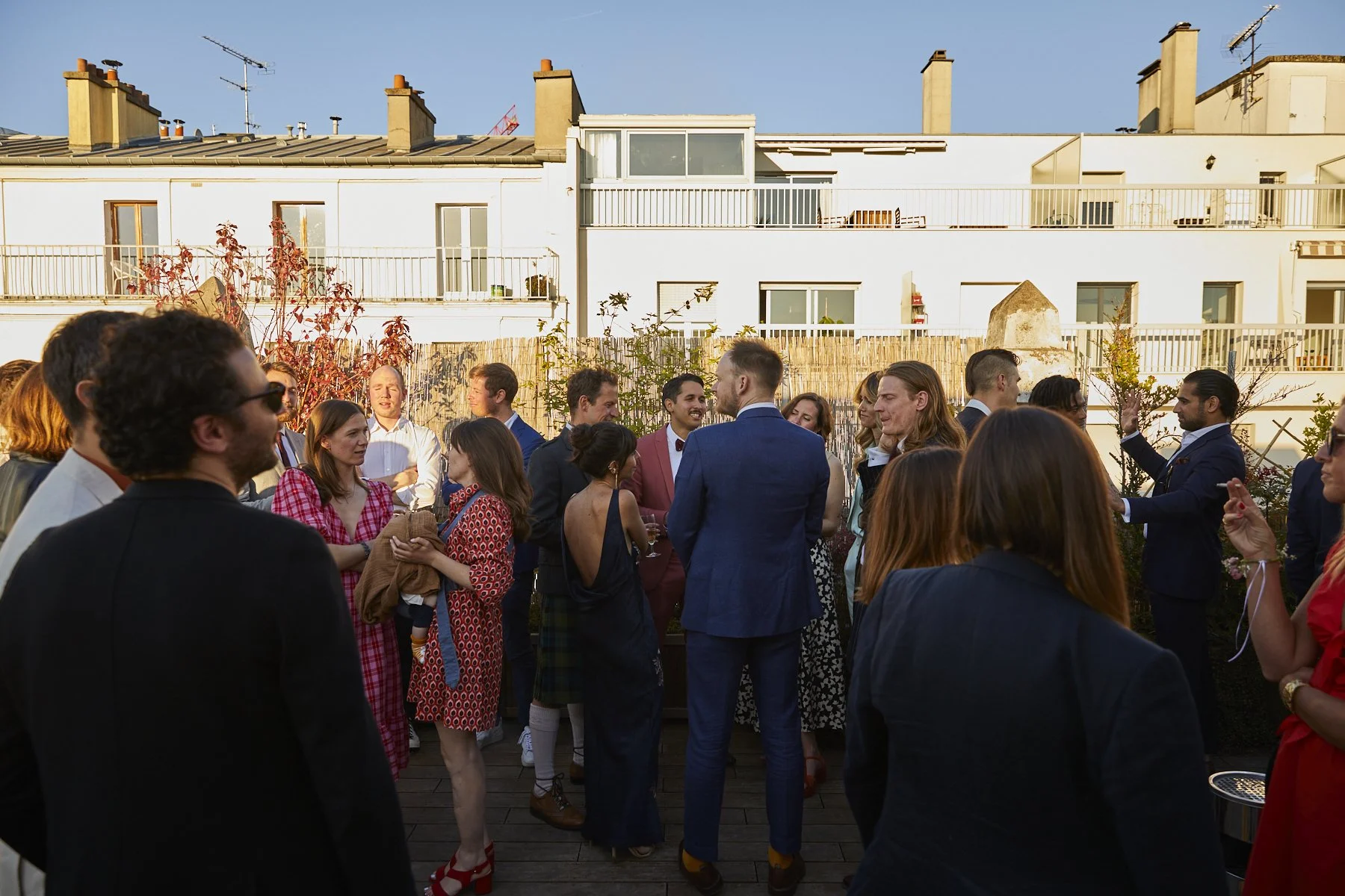 Group of people socializing at an outdoor rooftop party during daytime, with modern white buildings in the background.