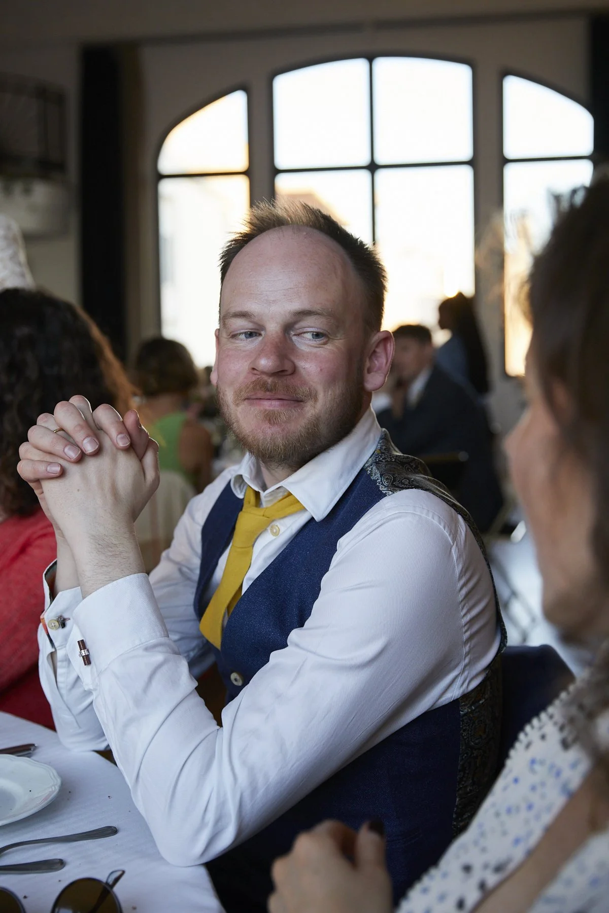 A man in a white shirt, yellow tie, and blue vest sitting at a dining table, with people in the background during a social gathering or event indoors near large windows.