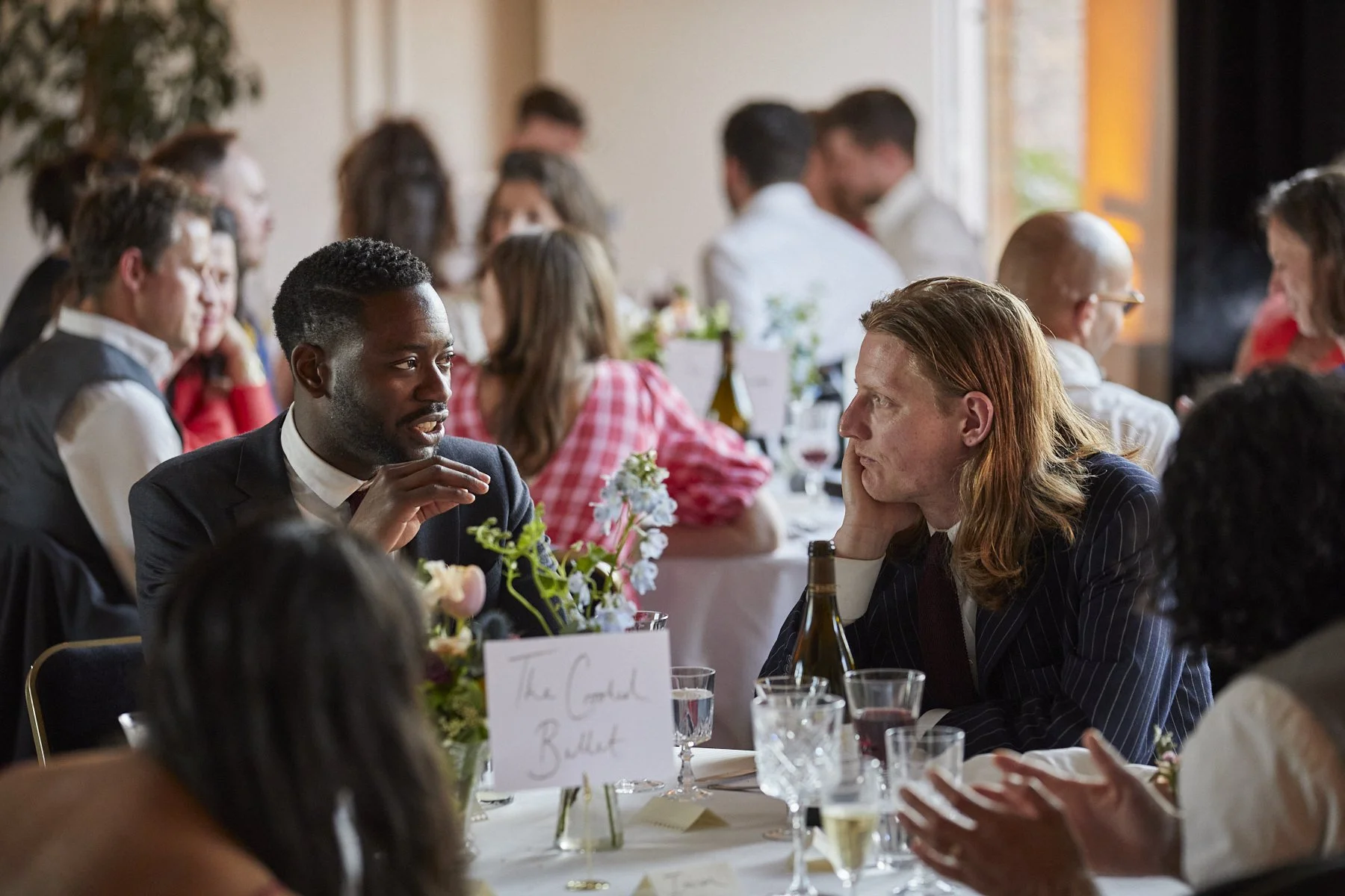 People seated at a banquet table engaged in conversation, with floral centerpieces and glasses of wine.