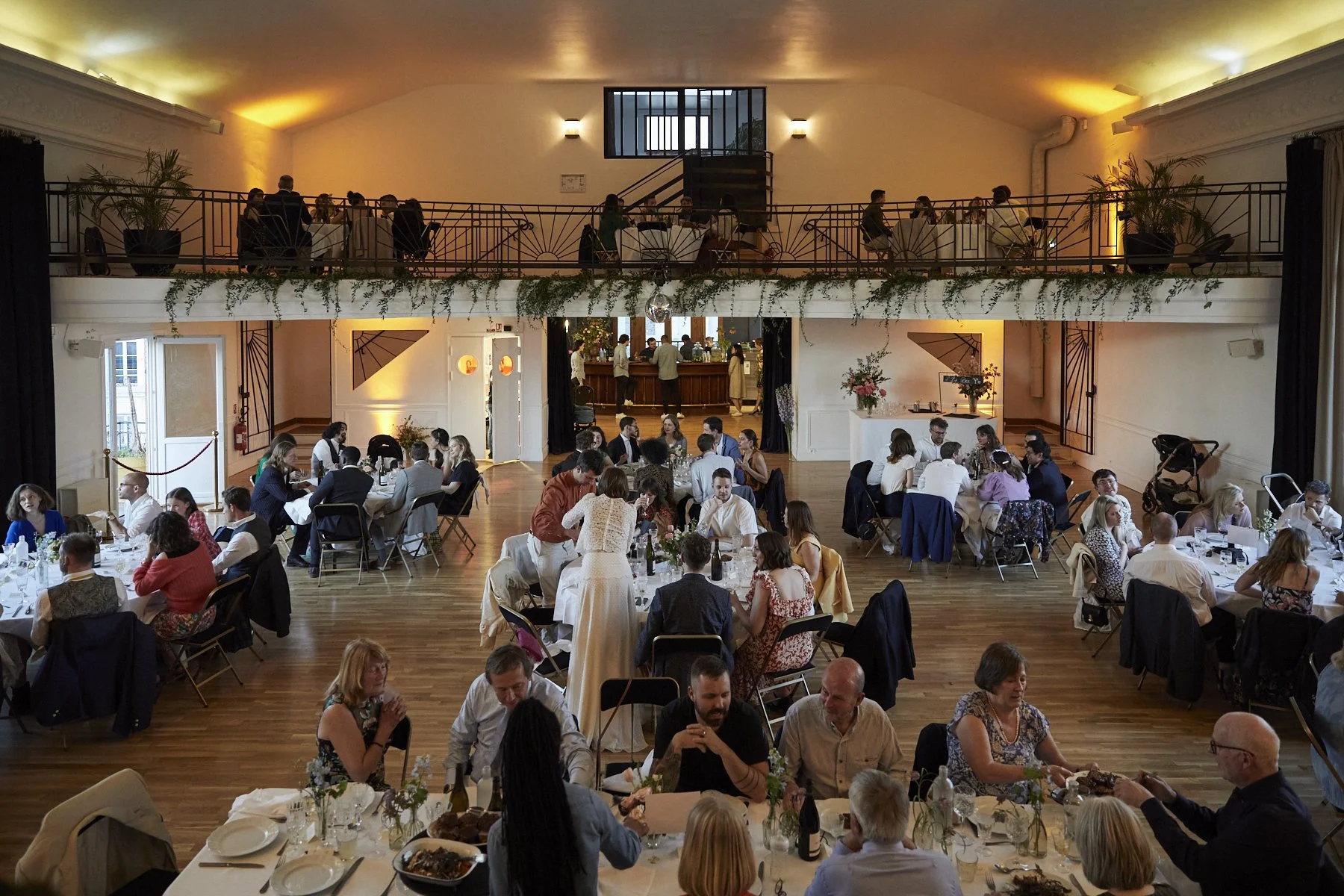 Indoor wedding reception with multiple round tables filled with guests, a dance floor, and a balcony with people dining, decorated with plants and warm lighting.