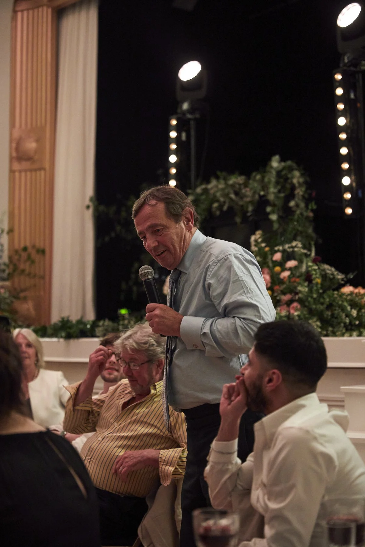 A man in a light blue shirt and tie is speaking into a microphone at an indoor event, surrounded by seated guests, with floral arrangements and stage lighting in the background.