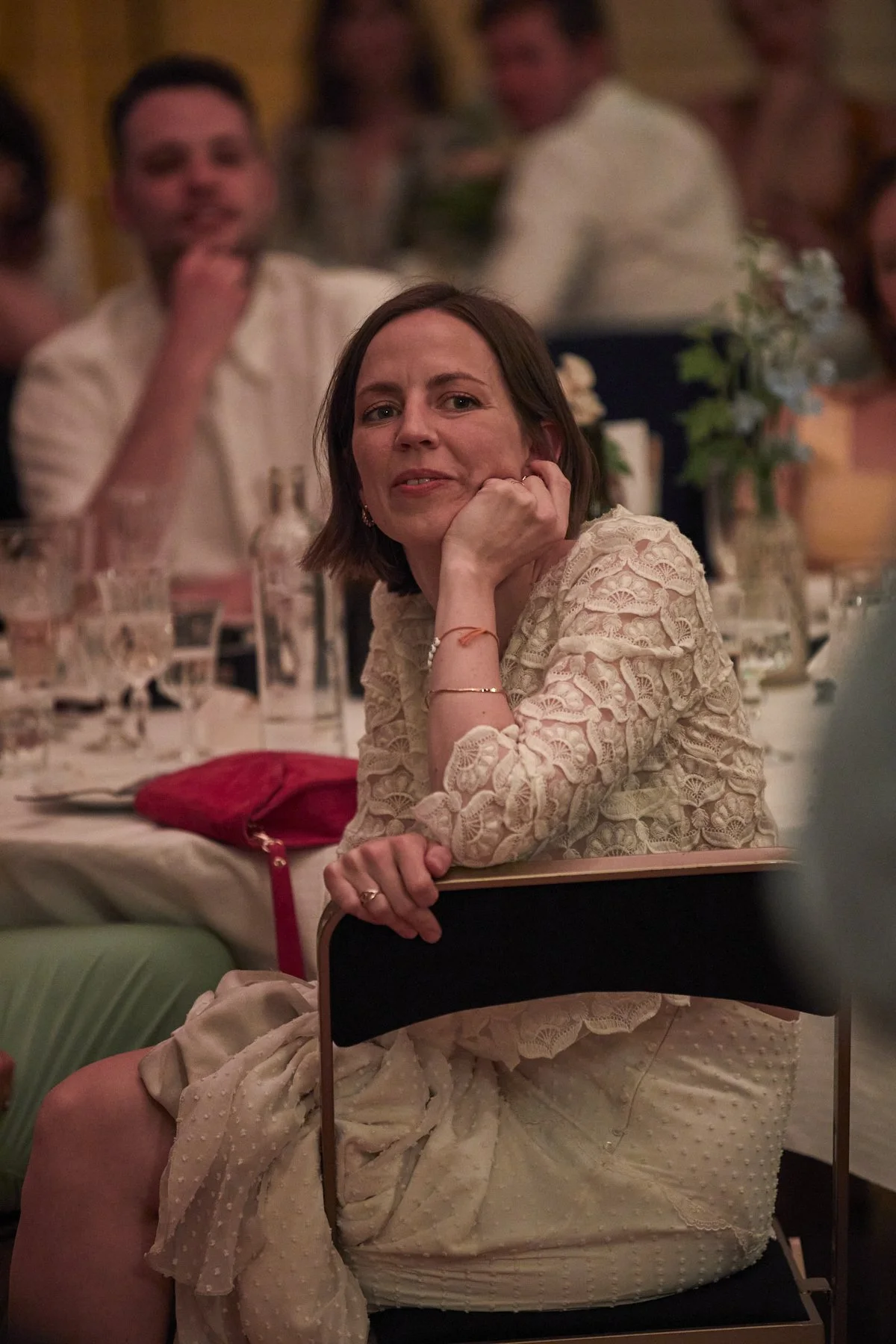 A woman sitting at a table with her hand resting on her chin, wearing a lace dress, at a formal event surrounded by other guests, with a man in the background.