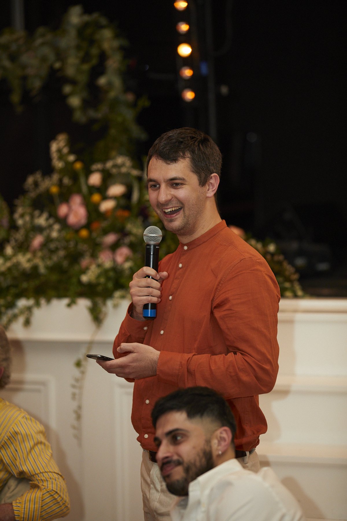 A man is standing, smiling, and holding a microphone and a smartphone, while speaking at an event. He has short dark hair and is wearing an orange button-up shirt. There are other people seated nearby and floral decorations in the background.