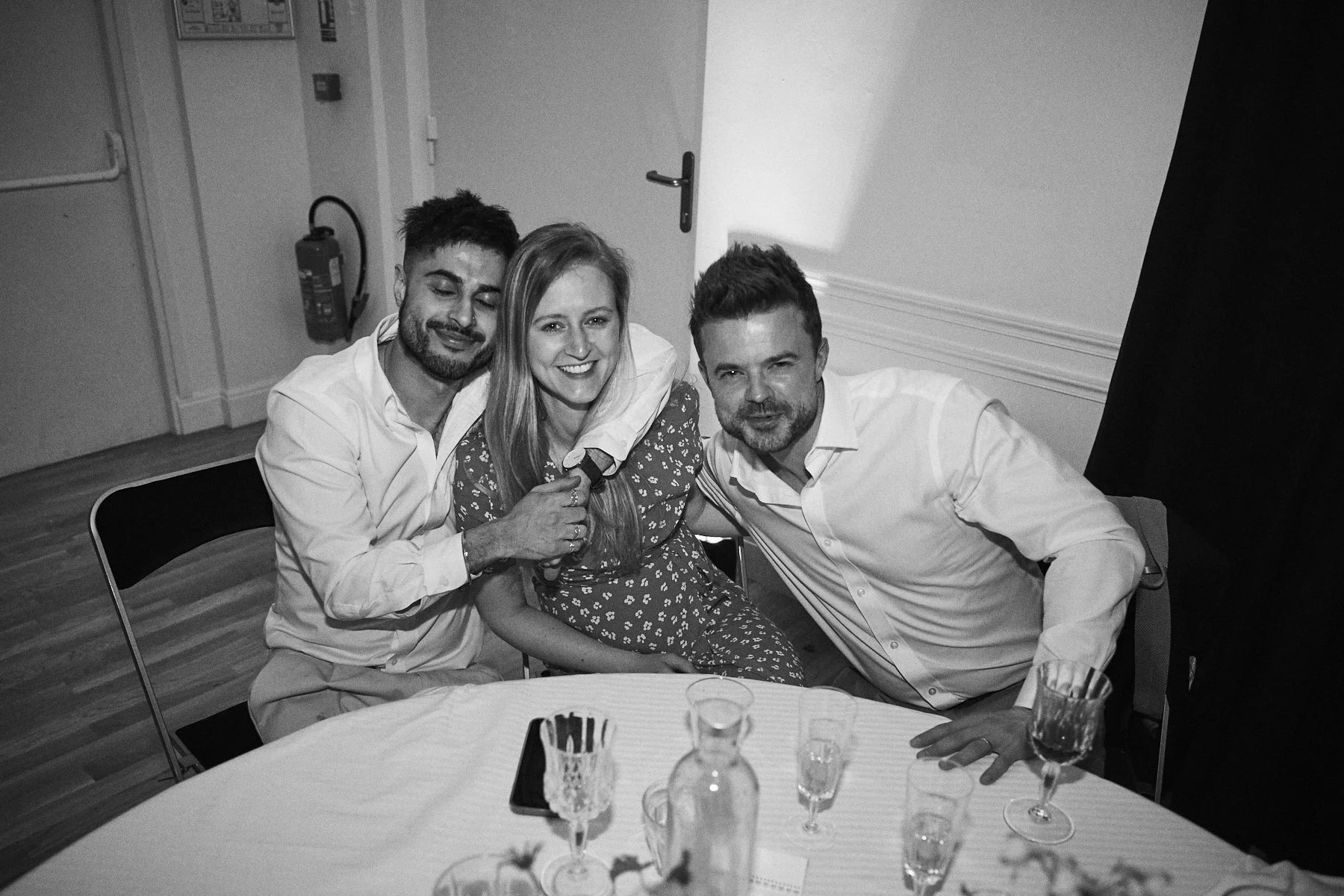 Three people sitting at a table, smiling and posing for the camera, with drinks and a phone on the table.