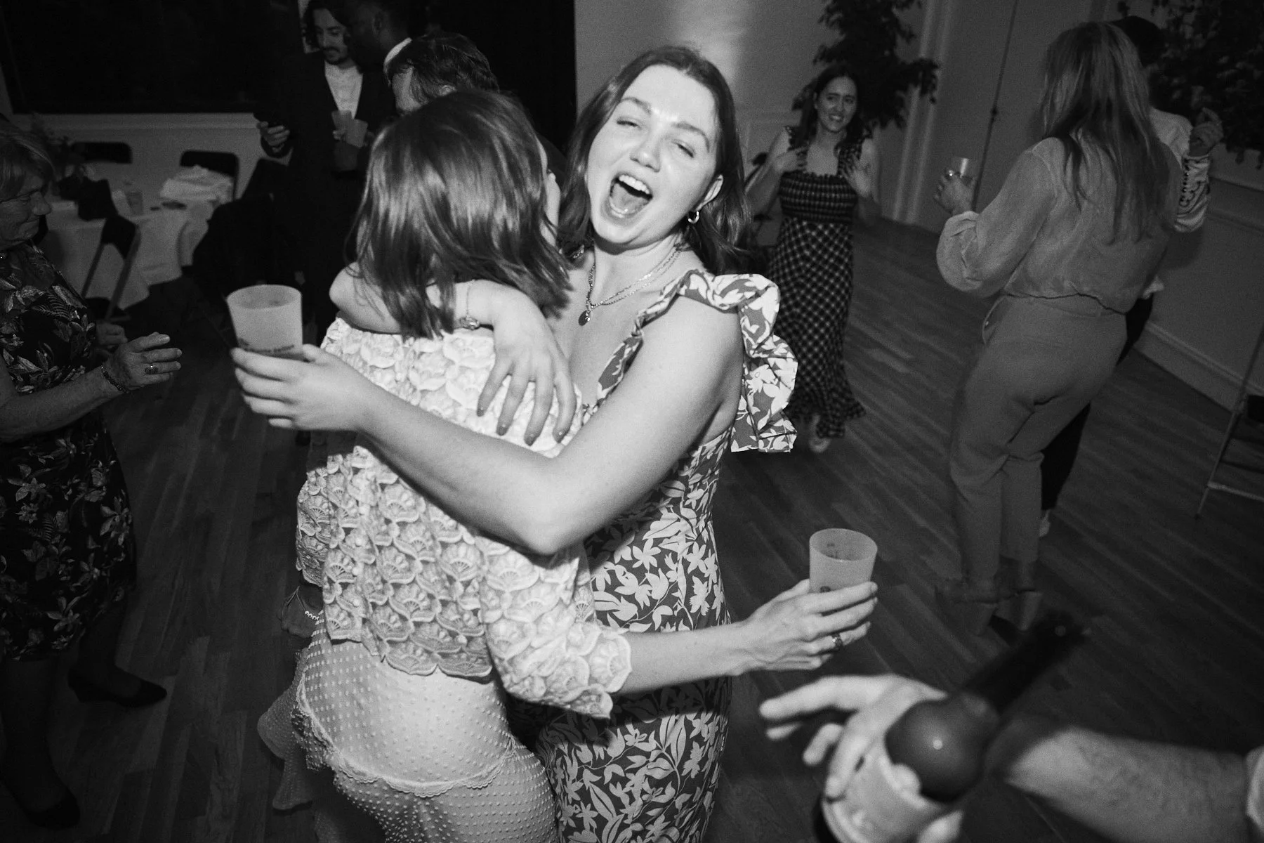 Two women are hugging and smiling at a celebration or party, with other people in the background dancing and holding drinks.