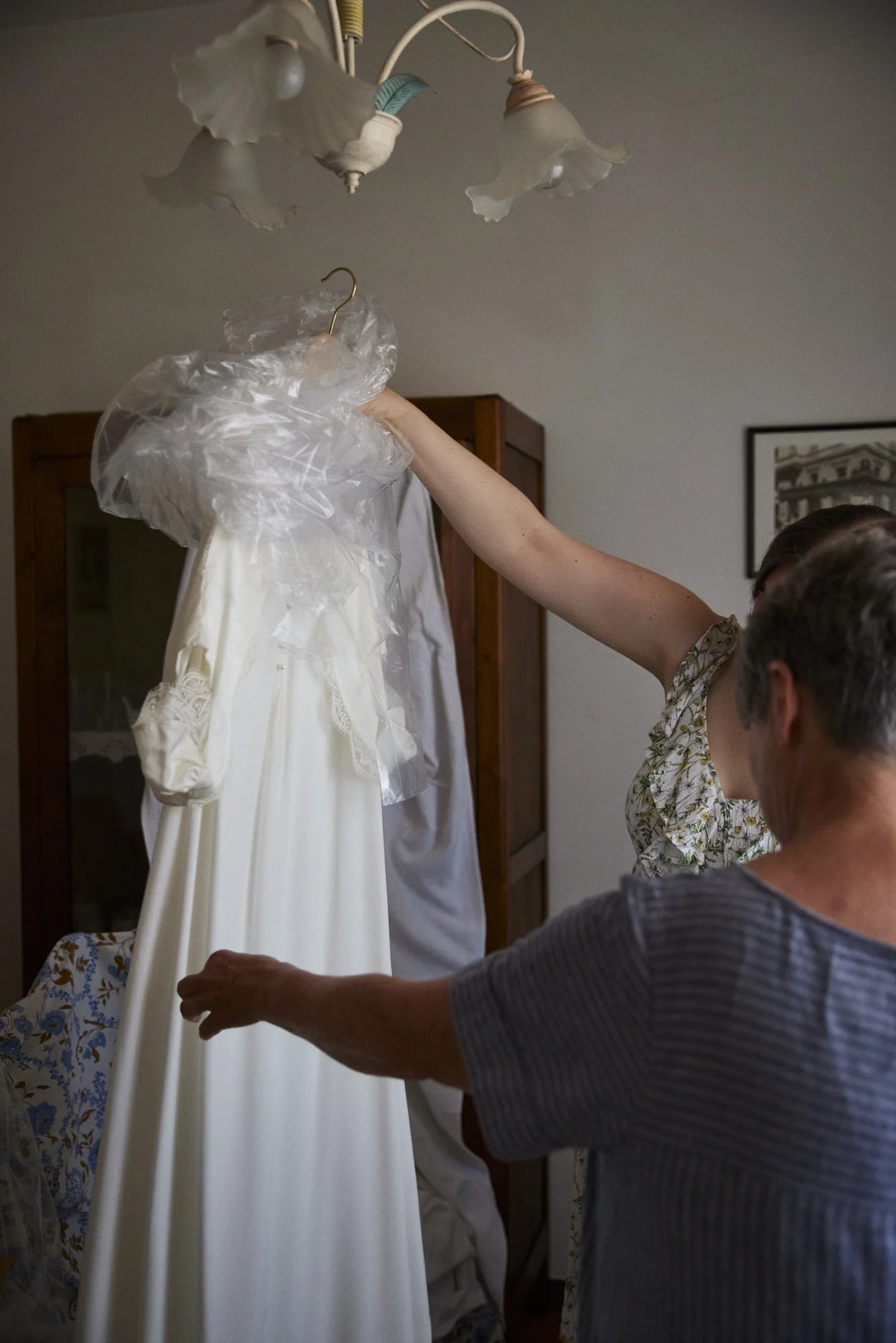 Two women are preparing a white wedding dress on a hanger in a room with a wooden wardrobe and a framed picture on the wall.