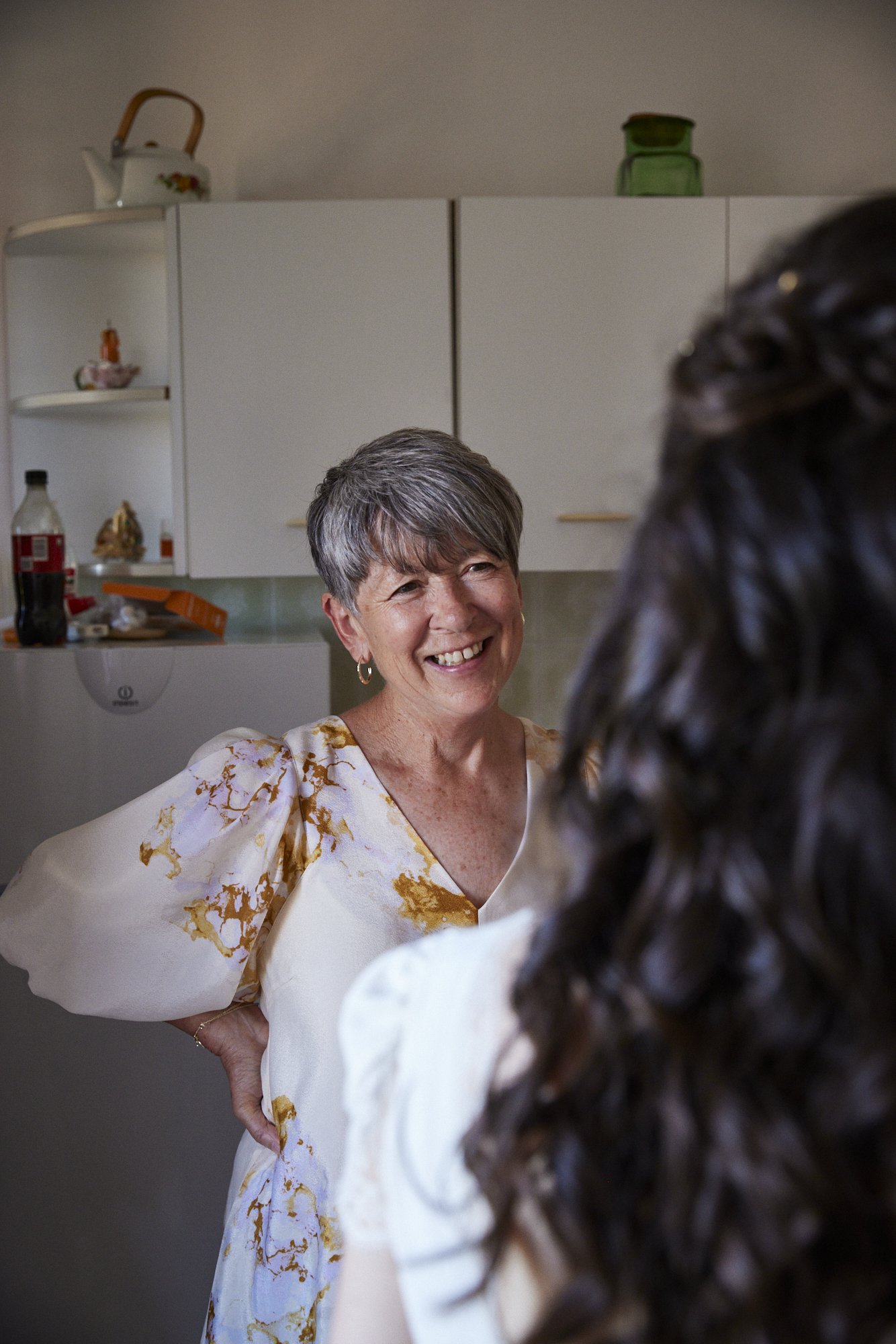 Smiling woman with short gray hair talking to another woman with long dark curly hair in a kitchen.