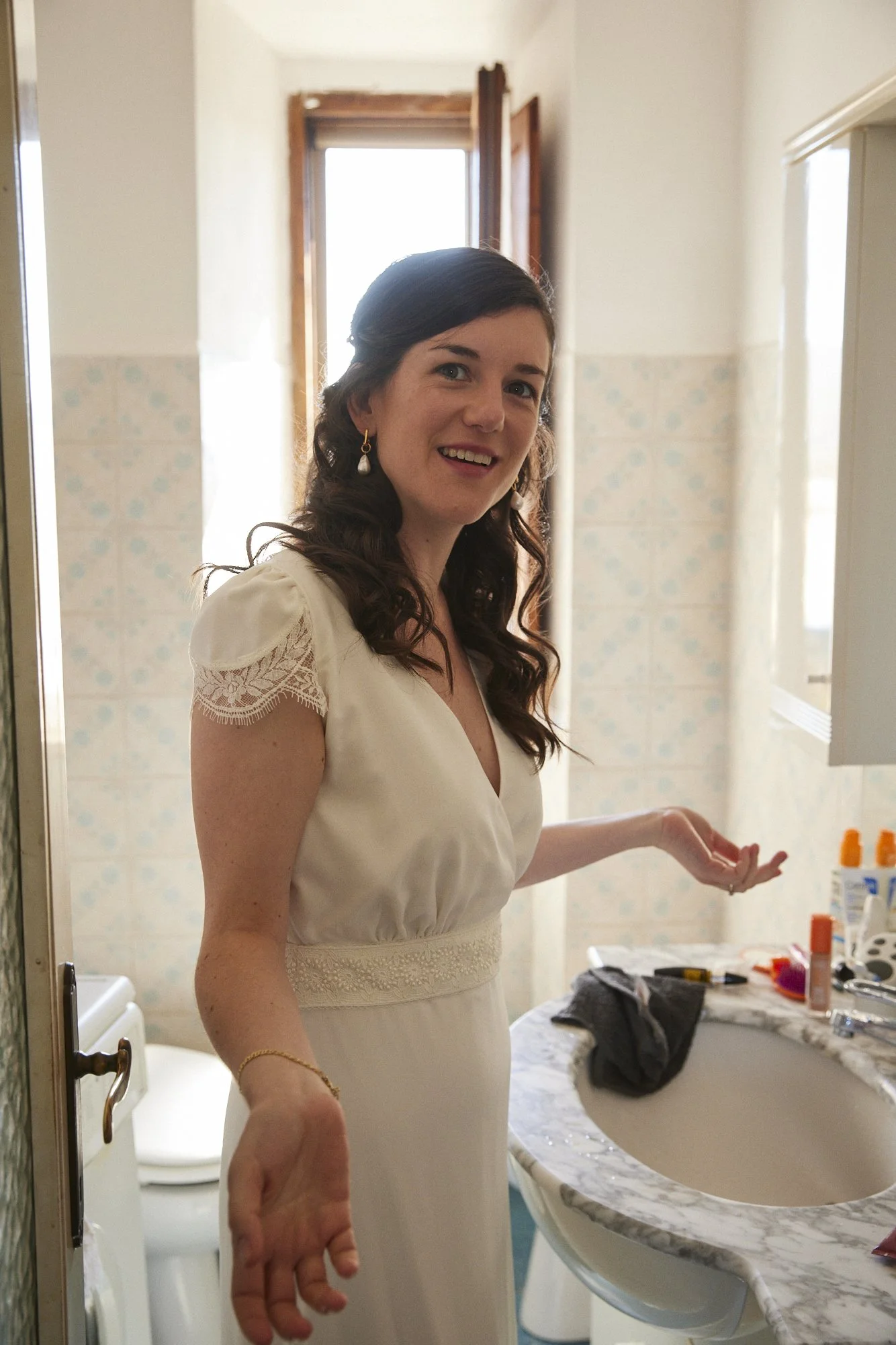 A young woman in a white dress stands in a bathroom, smiling at the camera, with her hands gesturing towards a marble countertop with toiletries and personal items.