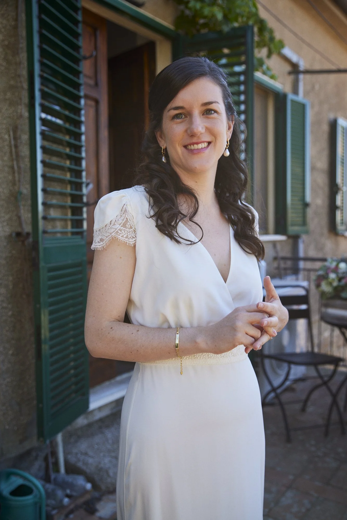 A woman with long dark curly hair, wearing a white dress with lace details on the sleeves, is smiling and standing outside near green window shutters and a rustic wall, with a patio area in the background.