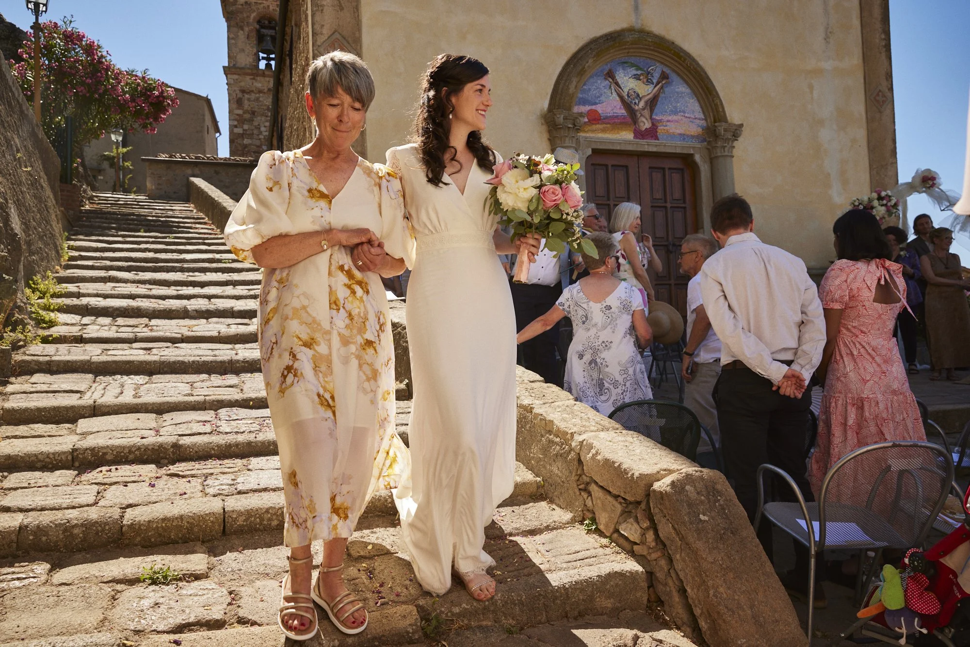 A bride in a white dress holding a bouquet walks down stone steps with a woman in a floral dress during a daytime outdoor wedding ceremony.