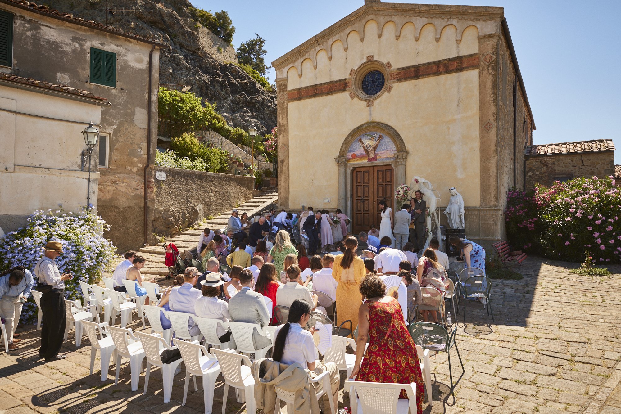 A wedding ceremony outdoors in front of a historic church, with guests seated on white chairs, some standing, and the officiant and couple near the church entrance, surrounded by flowering bushes in a sunny setting.