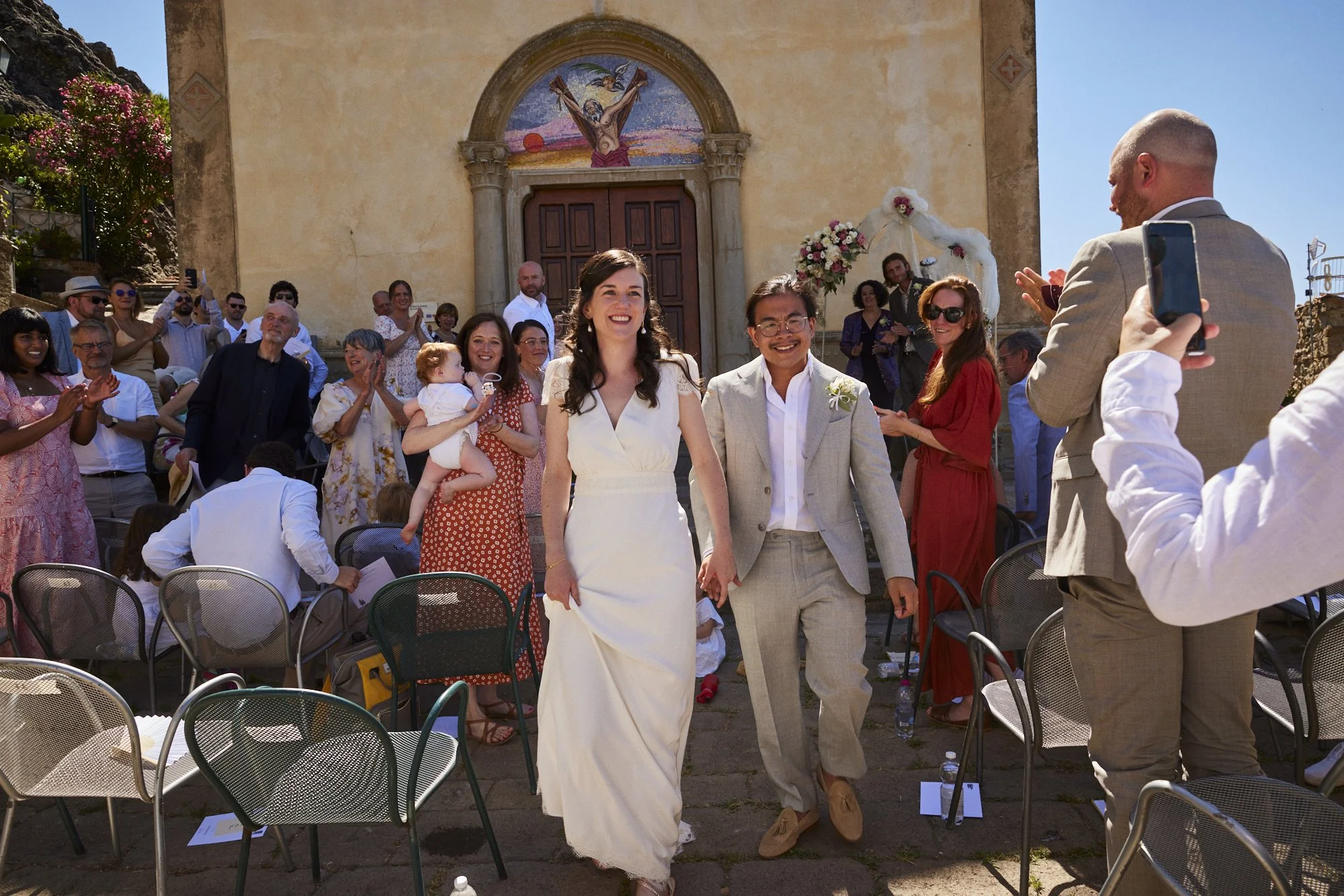 A newly married couple walking hand in hand down the aisle after a wedding ceremony outside a church with a mural of Jesus Christ on the wall behind them. They are smiling and surrounded by friends and family clapping and celebrating.