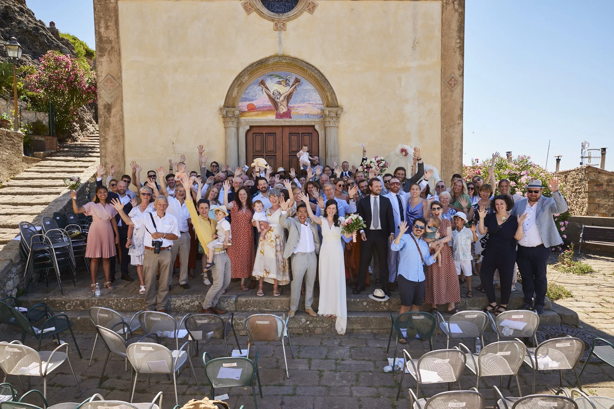 A large group of people gathered outside a church, celebrating a wedding day, with some waving at the camera, others holding flowers, and children present, under a sunny sky.