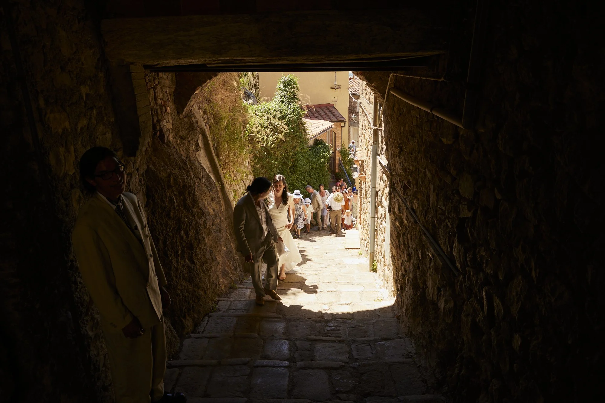 People walking up a stone alleyway under an overpass on a sunny day, with some people dressed in formal attire.