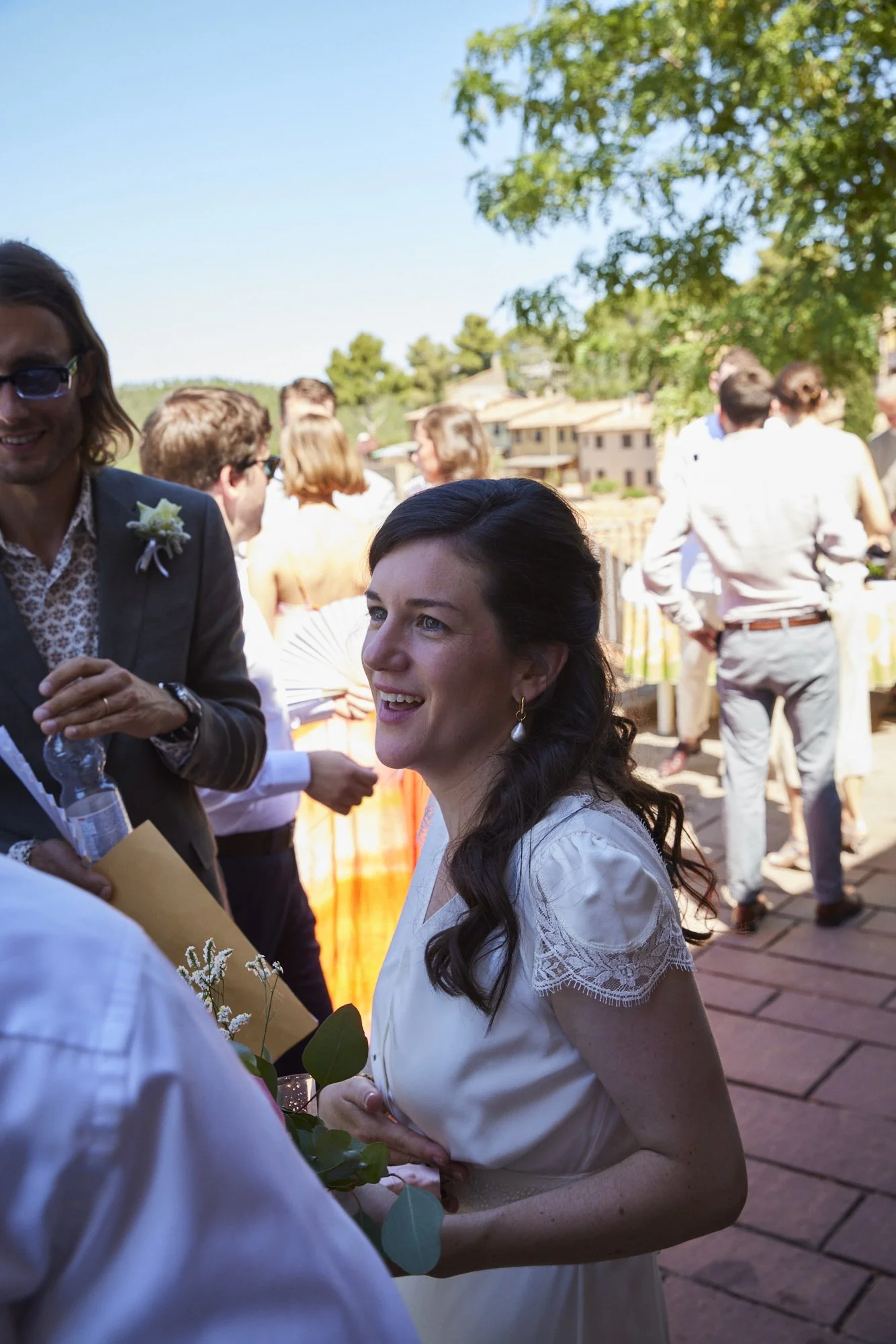 A woman with dark hair in a white dress smiling at an outdoor wedding reception. She holds a small bouquet of flowers, and there are guests and a scenic outdoor background with trees and buildings.