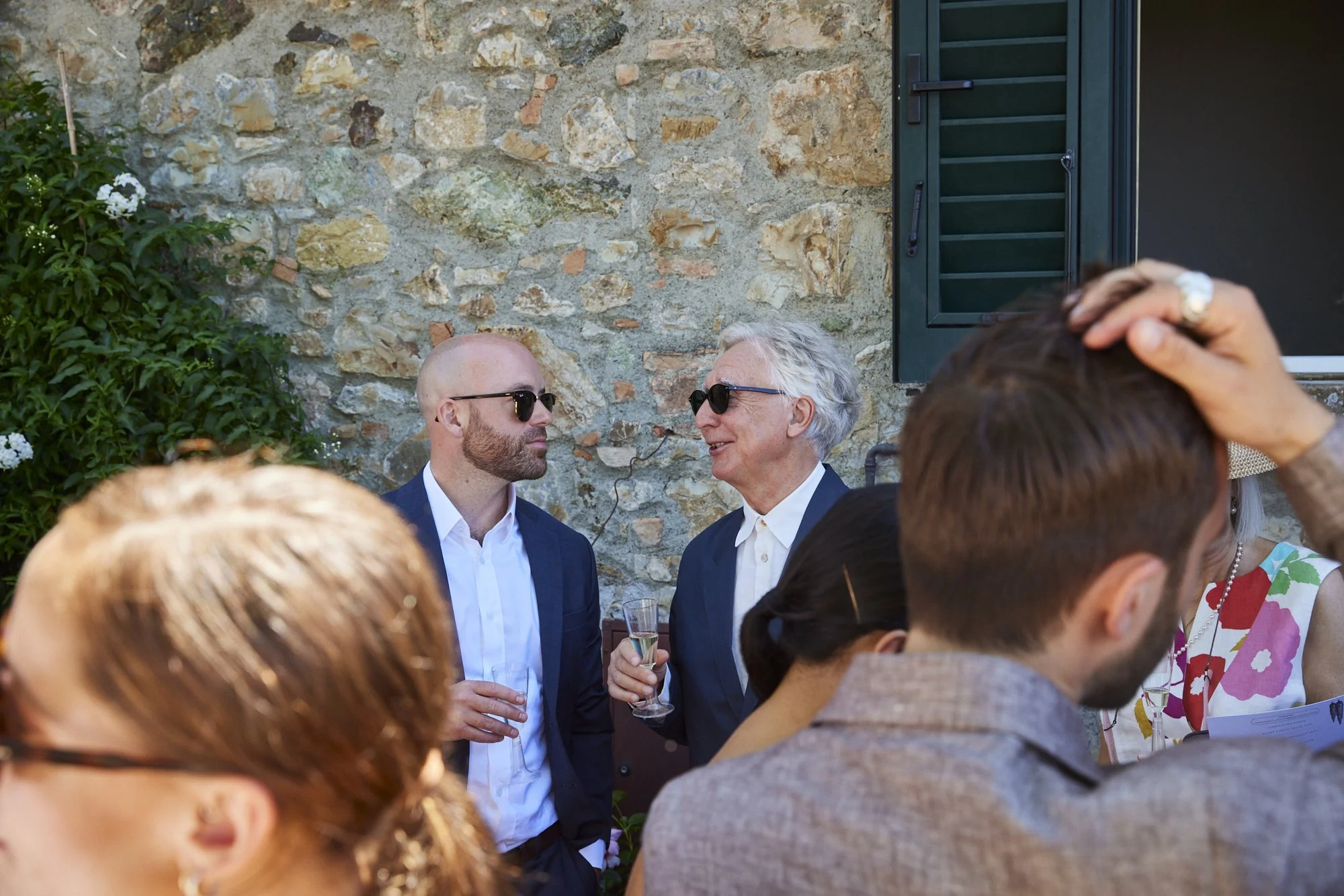 Two men with sunglasses, one older with gray hair and one younger with a bald head, are talking at an outdoor gathering near a stone wall. The older man is holding a glass of champagne, and other guests are visible in the foreground and background.