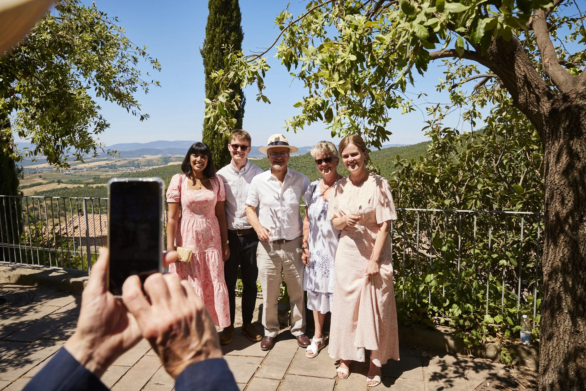 Group of six people in summer clothes taking a photo outdoors under trees with landscape background.