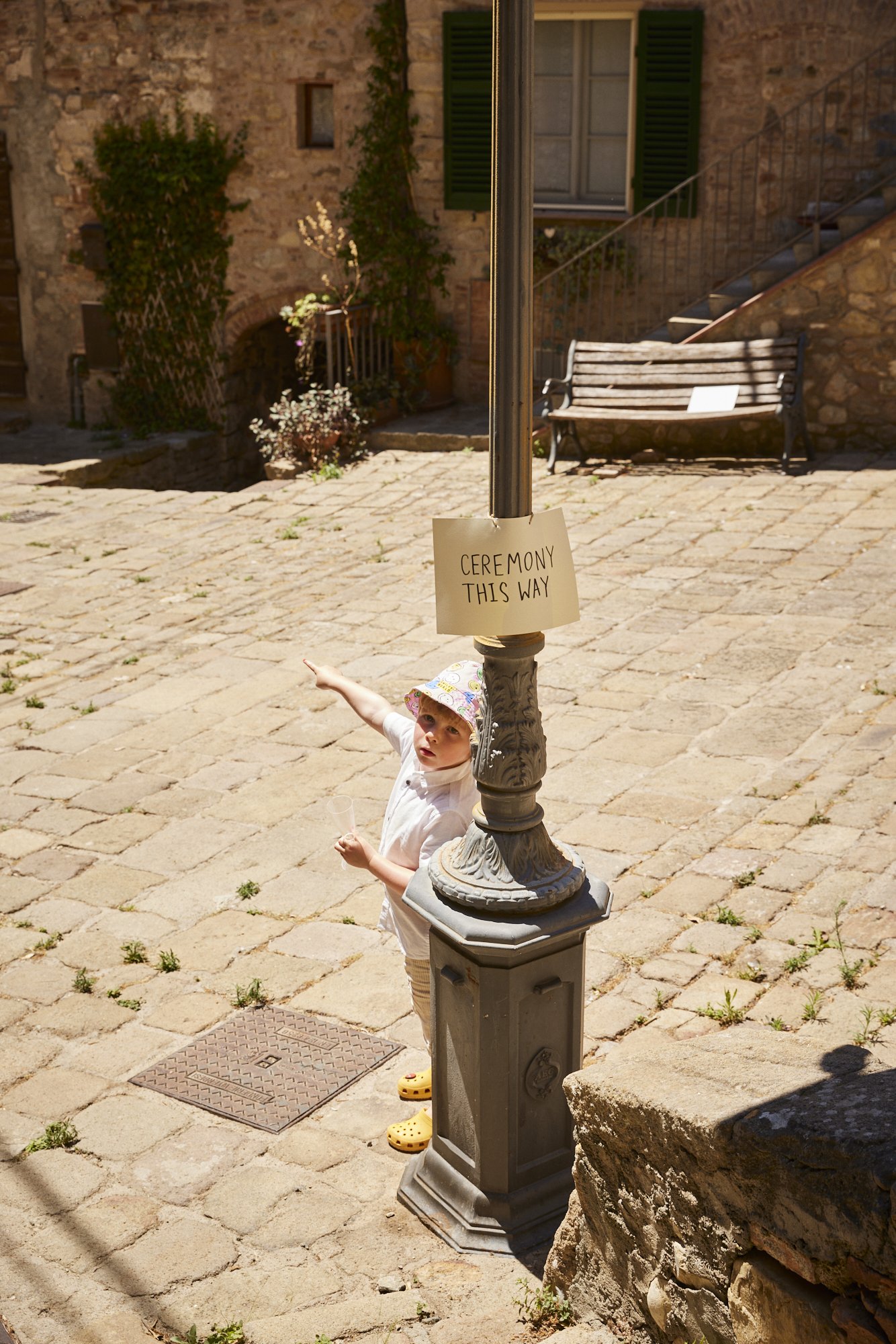 A young child wearing a colorful hat, white shirt, and yellow Crocs is standing near a lamppost with a sign that says 'Ceremony This Way.' The child appears to be pointing or directing. The setting is an outdoor courtyard with stone pavement and a st