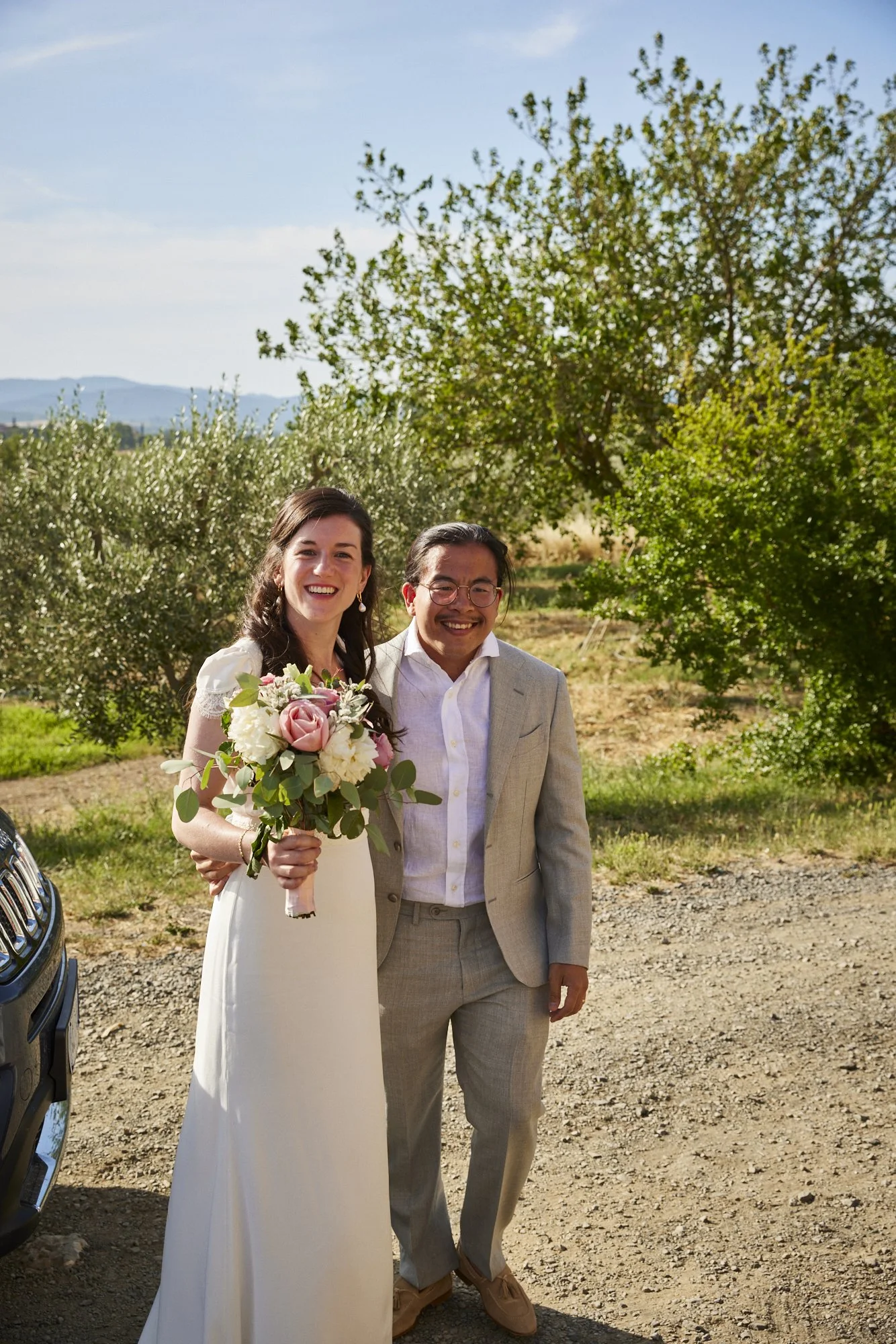 A smiling bride in a white dress holding a bouquet of pink and white flowers, standing next to a smiling groom in a light grey suit, outdoors with green trees and blue sky in the background.