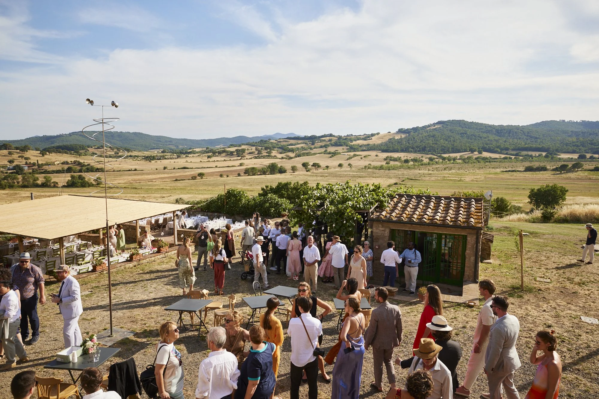 Guests at an outdoor wedding reception in a rural field, with tables, chairs, and a small building, surrounded by rolling hills and farmland.