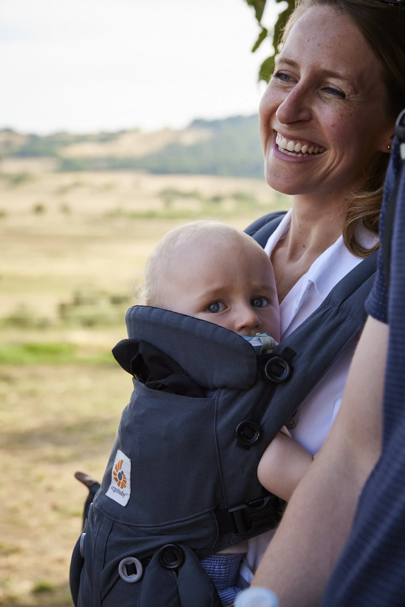 A woman with red hair and freckles is smiling while carrying a baby in a black baby carrier outdoors. The baby has light hair and blue eyes, and is looking over the carrier with a curious expression. The background features a blurred landscape of rol