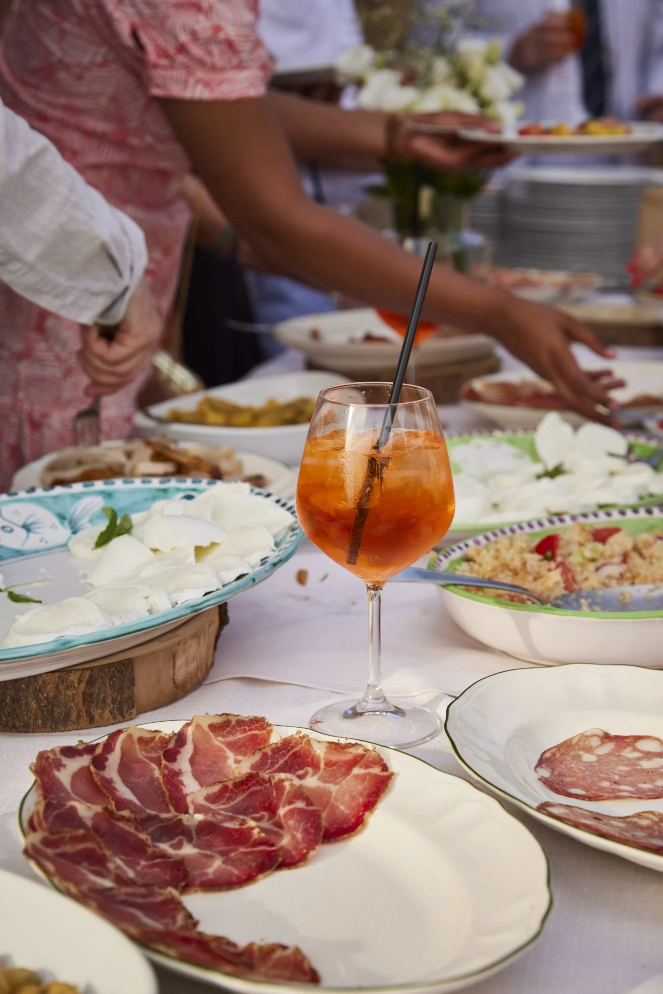 A table set with various plates of food including slices of cured meat, cheese, and bread, along with a glass of orange-colored drink with a straw. People are gathered around, serving themselves dishes at a buffet-style meal.