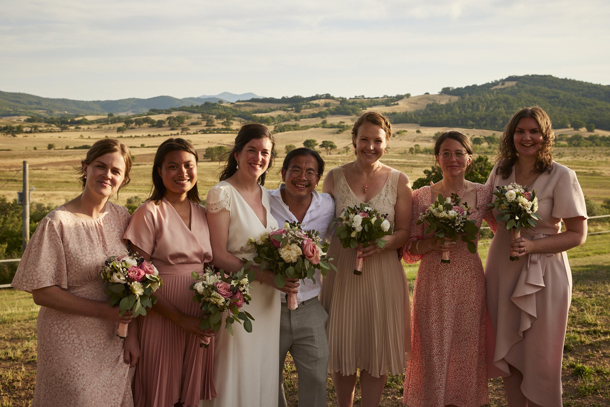 Group of seven people, including six women and one man, dressed in pastel-colored dresses and suits, holding bouquets of flowers, standing outdoors with rolling hills and a partly cloudy sky in the background.
