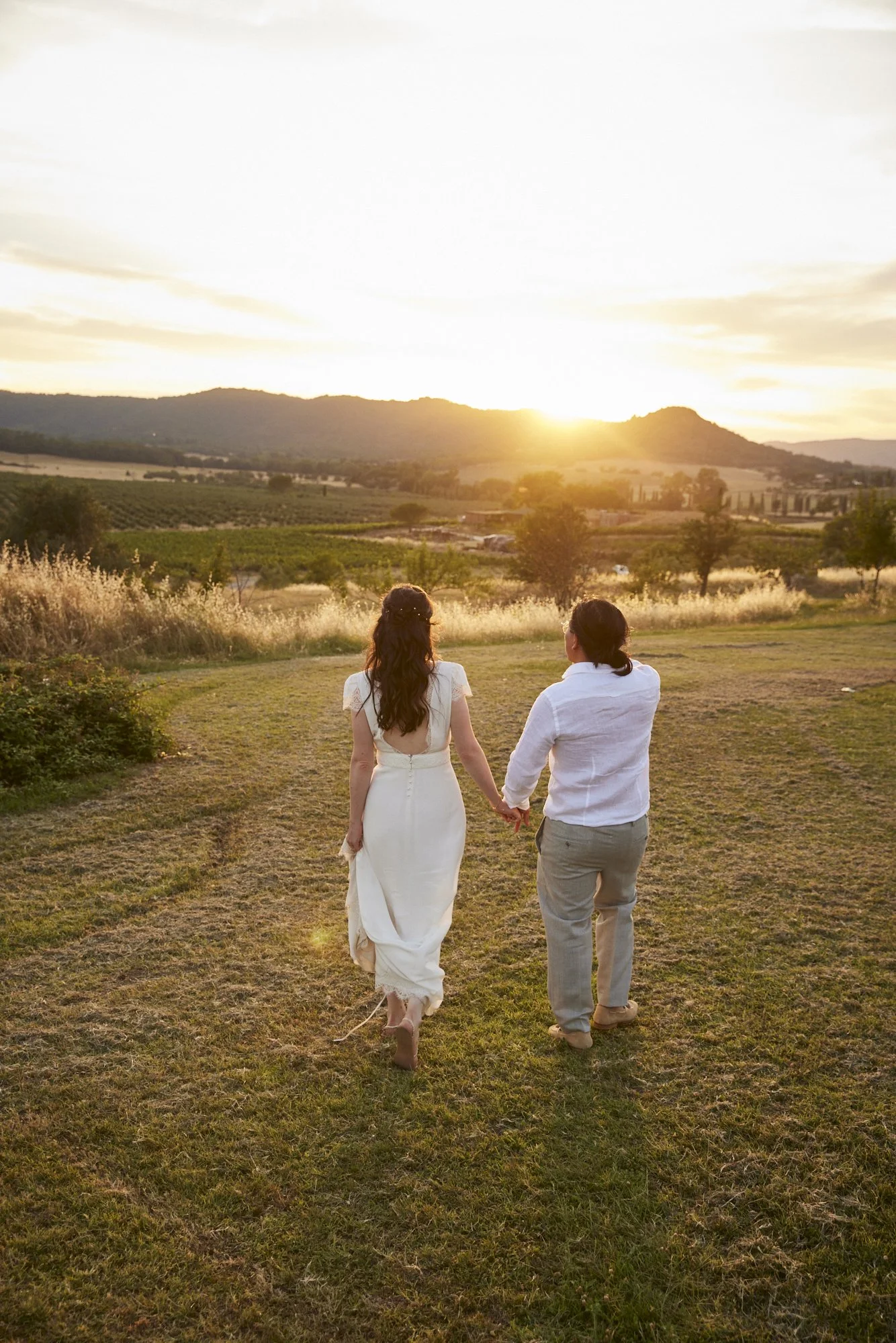 A couple holding hands and walking away on a grassy field at sunset, with a scenic landscape of hills and fields in the background.