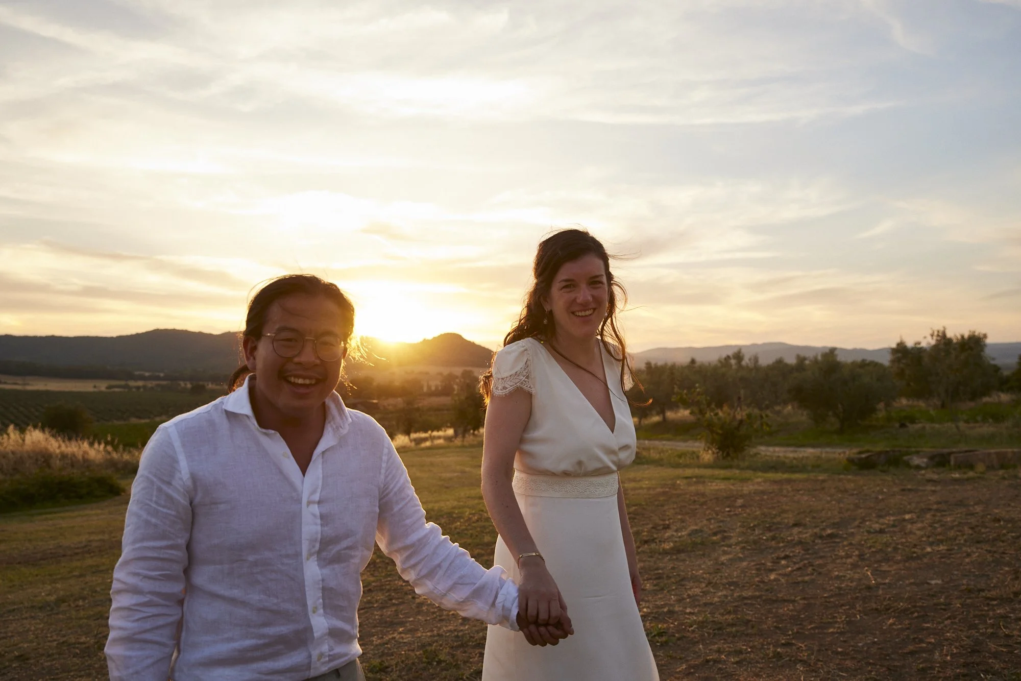 A smiling couple holding hands outdoors during sunset, with a scenic landscape and mountains in the background.