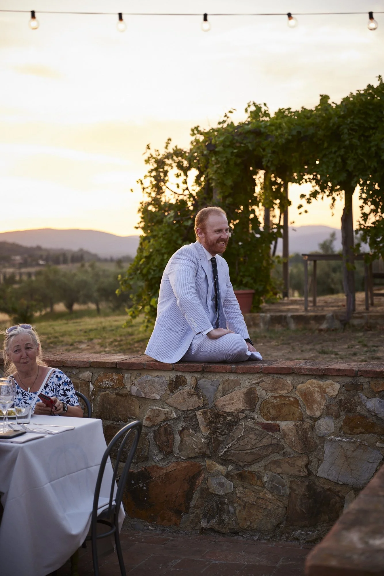 A man in a light-colored suit sitting on a stone wall outdoors during sunset, smiling, with a woman nearby at a dining table.