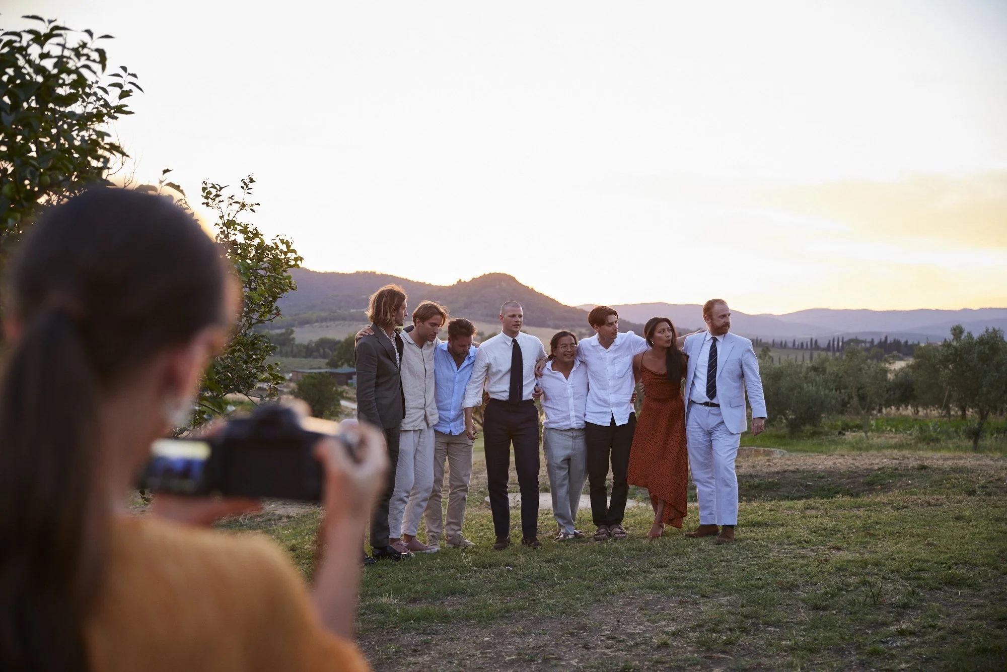 Group of eight people standing outdoors, embracing, during sunset, in a scenic rural landscape.