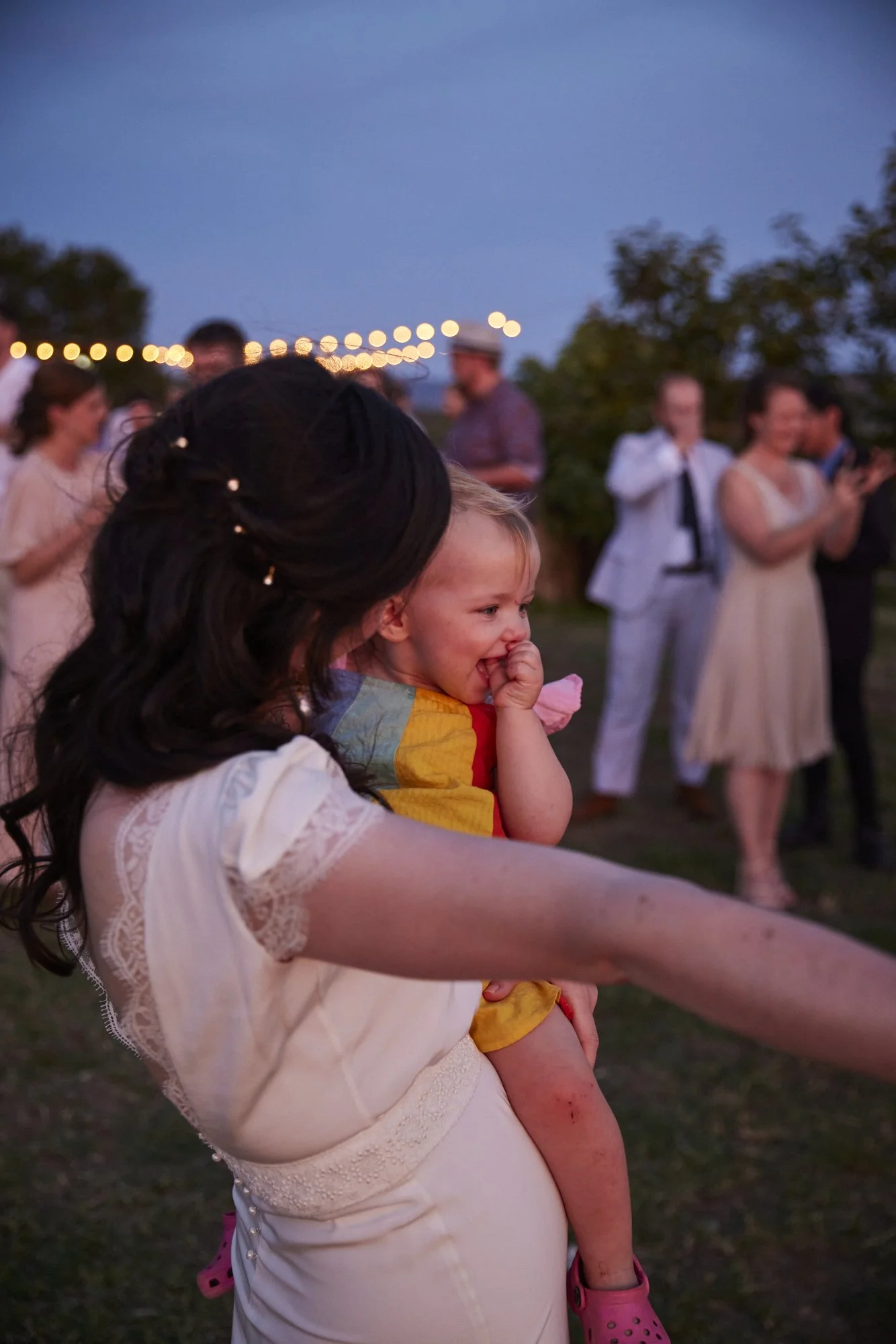 A woman holding a young girl at an outdoor gathering during dusk, with string lights and several people in the background.