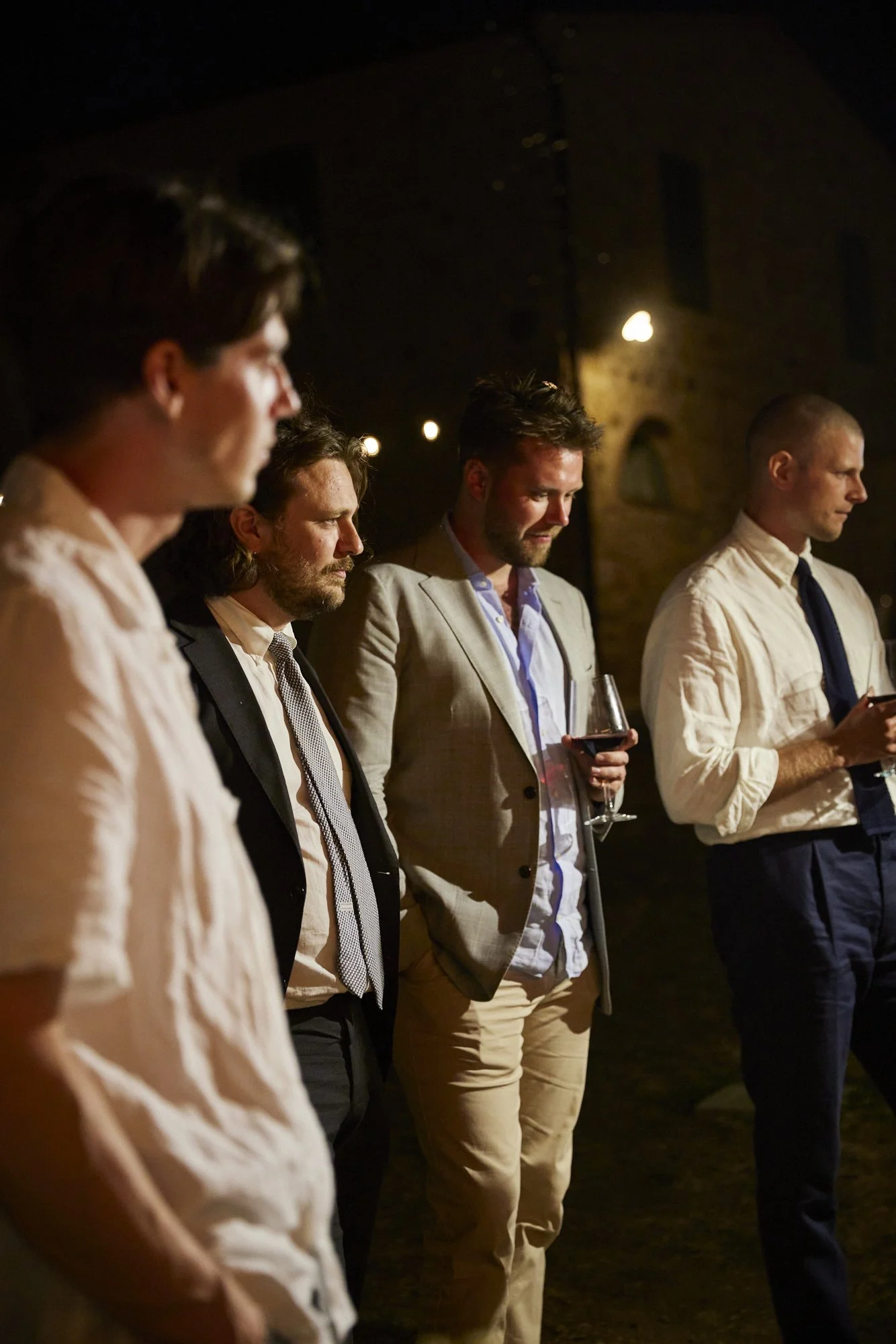 Five men standing in a row at an outdoor nighttime event, dressed in semi-formal attire, with some holding glasses of red wine, illuminated by warm lighting.