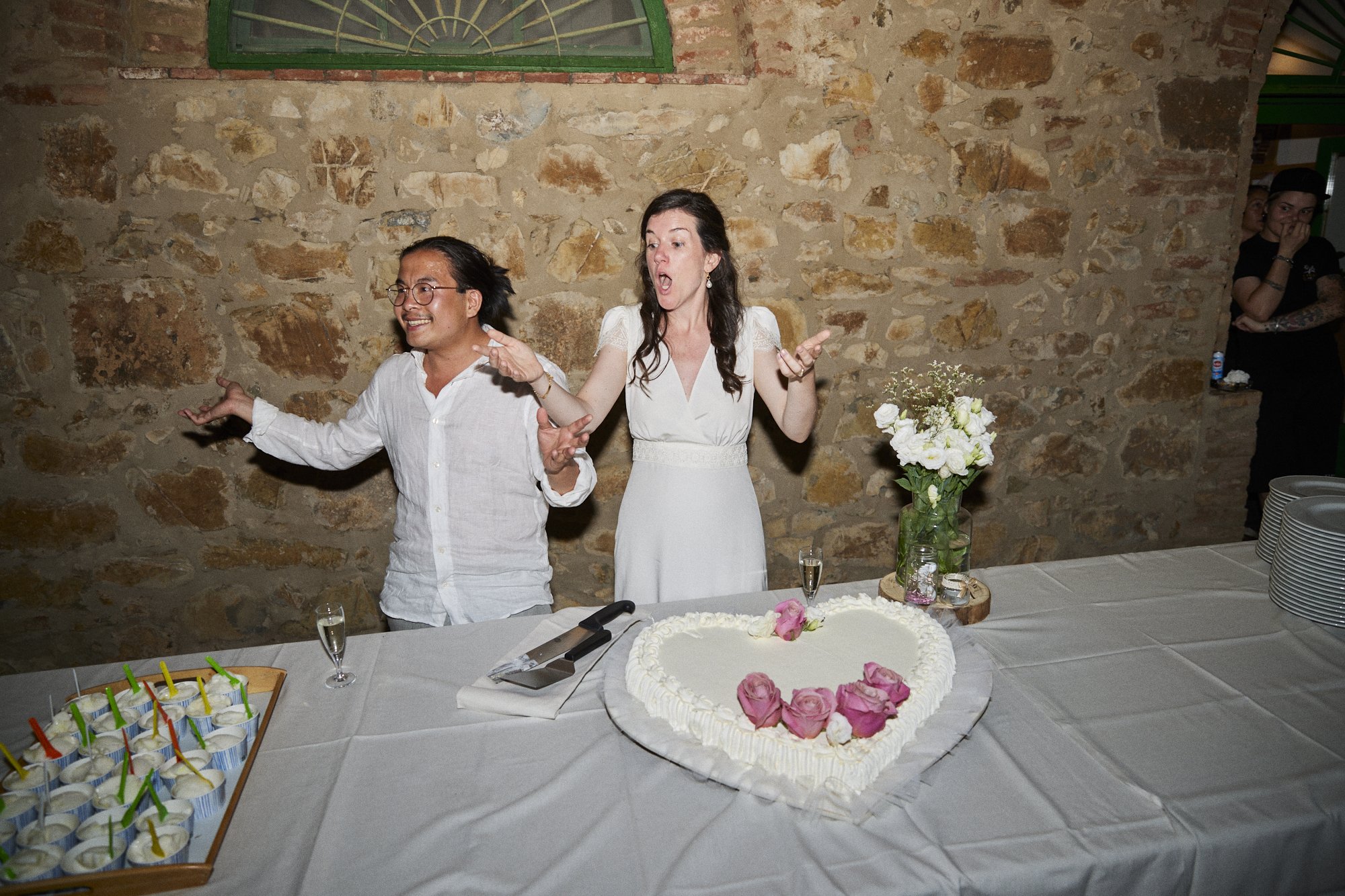 People celebrating a wedding cake, two individuals are in the center, one wearing glasses and a white shirt, the other in a white dress, with a heart-shaped cake decorated with pink roses on their table.