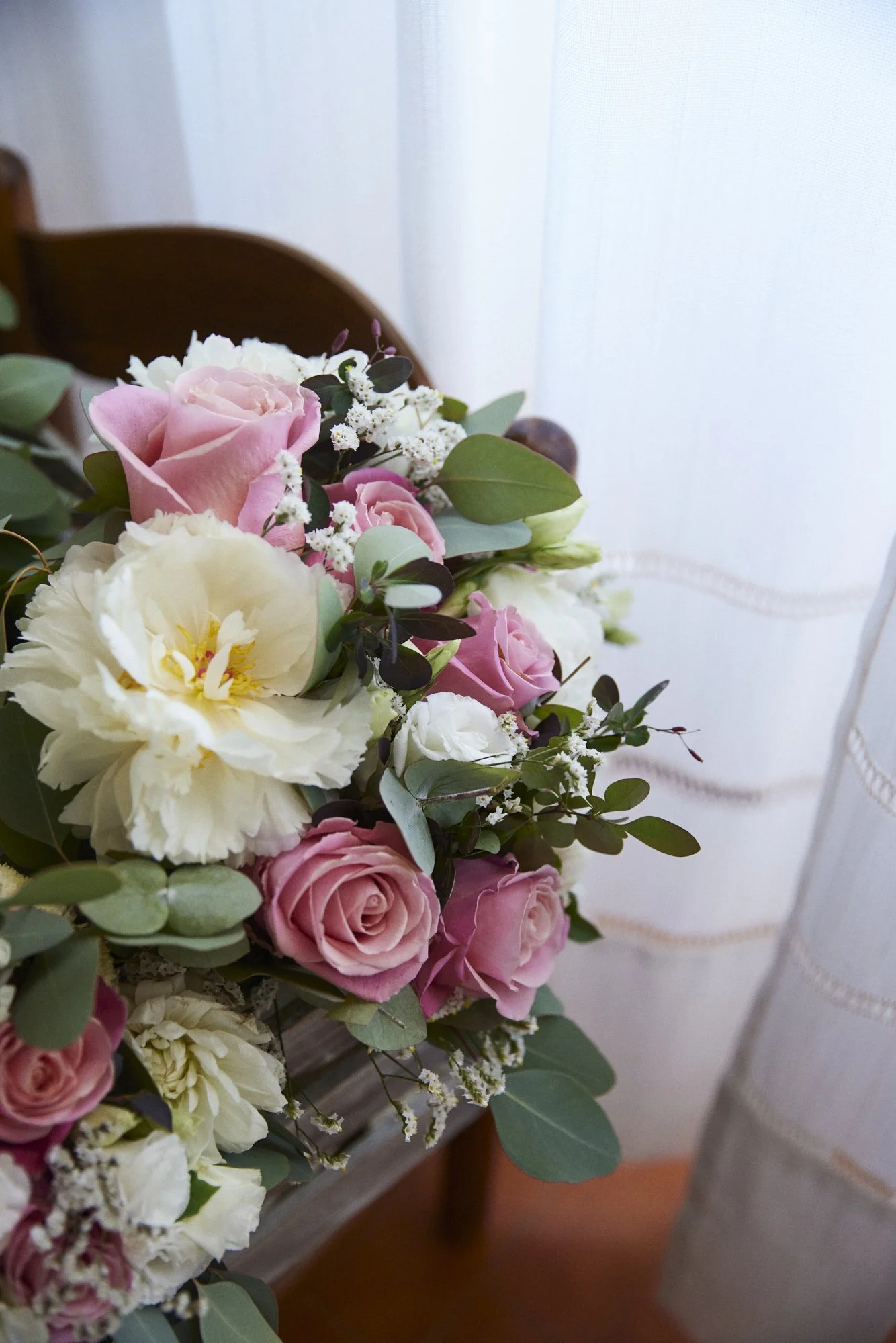 A close-up of a bouquet of pink roses, white carnations, and small white filler flowers with green leaves, placed on a wooden chair near a window with white curtains.